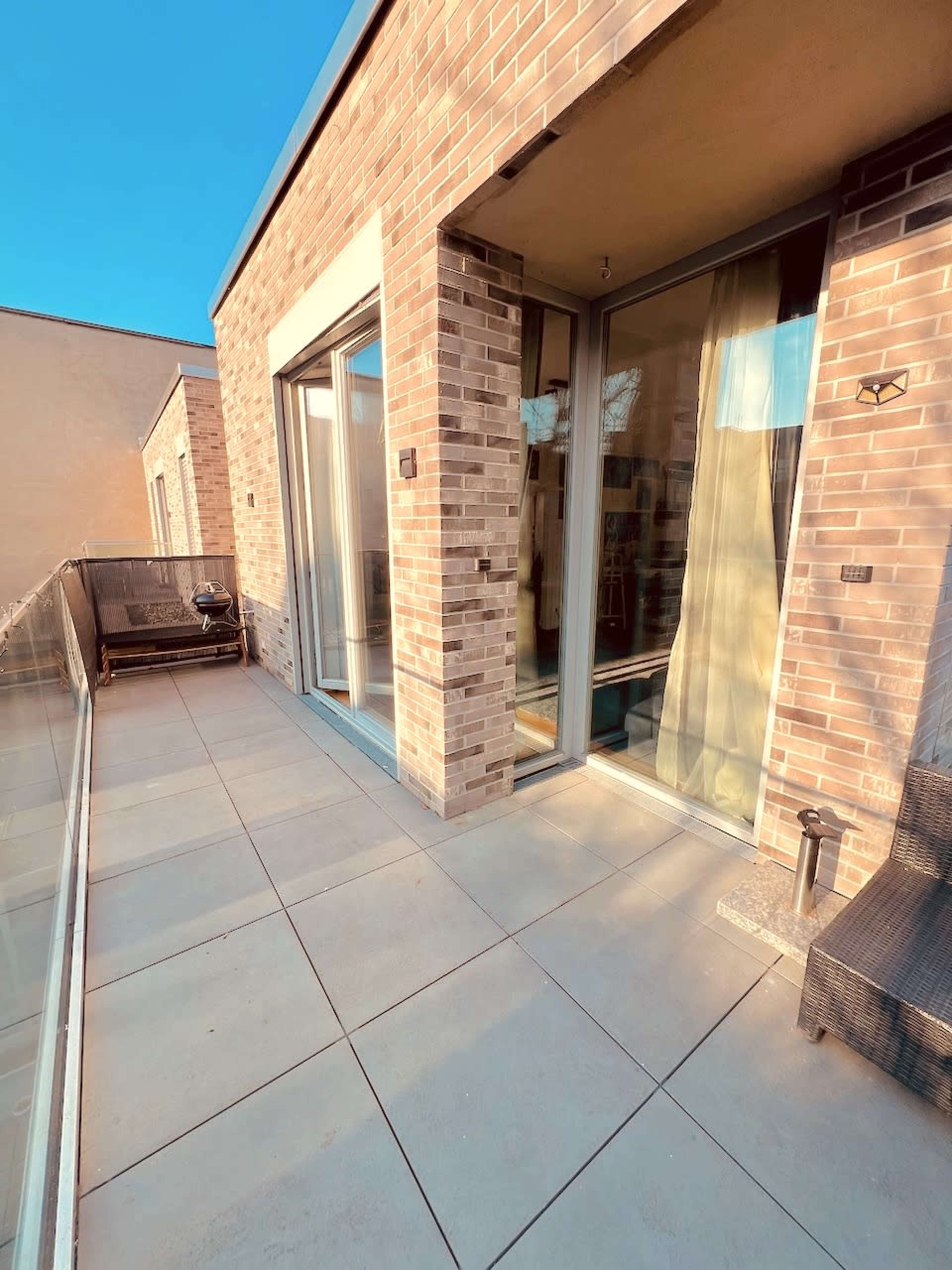 The image shows a modern balcony with large glass doors, brick walls, and a tiled floor, featuring a small seating area.