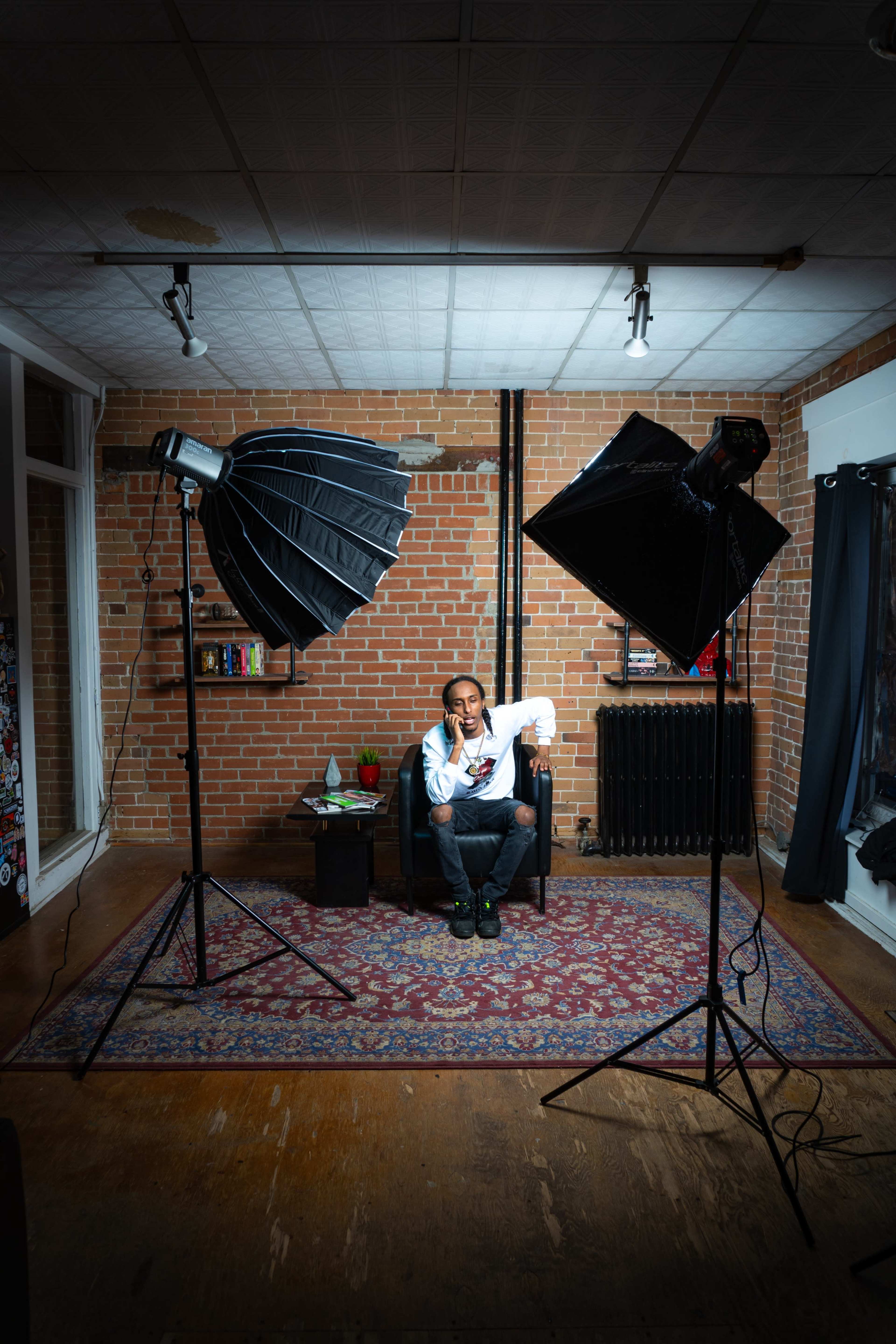A person sits on a chair in a room with brick walls, surrounded by two softbox lights and a small table with a plant.