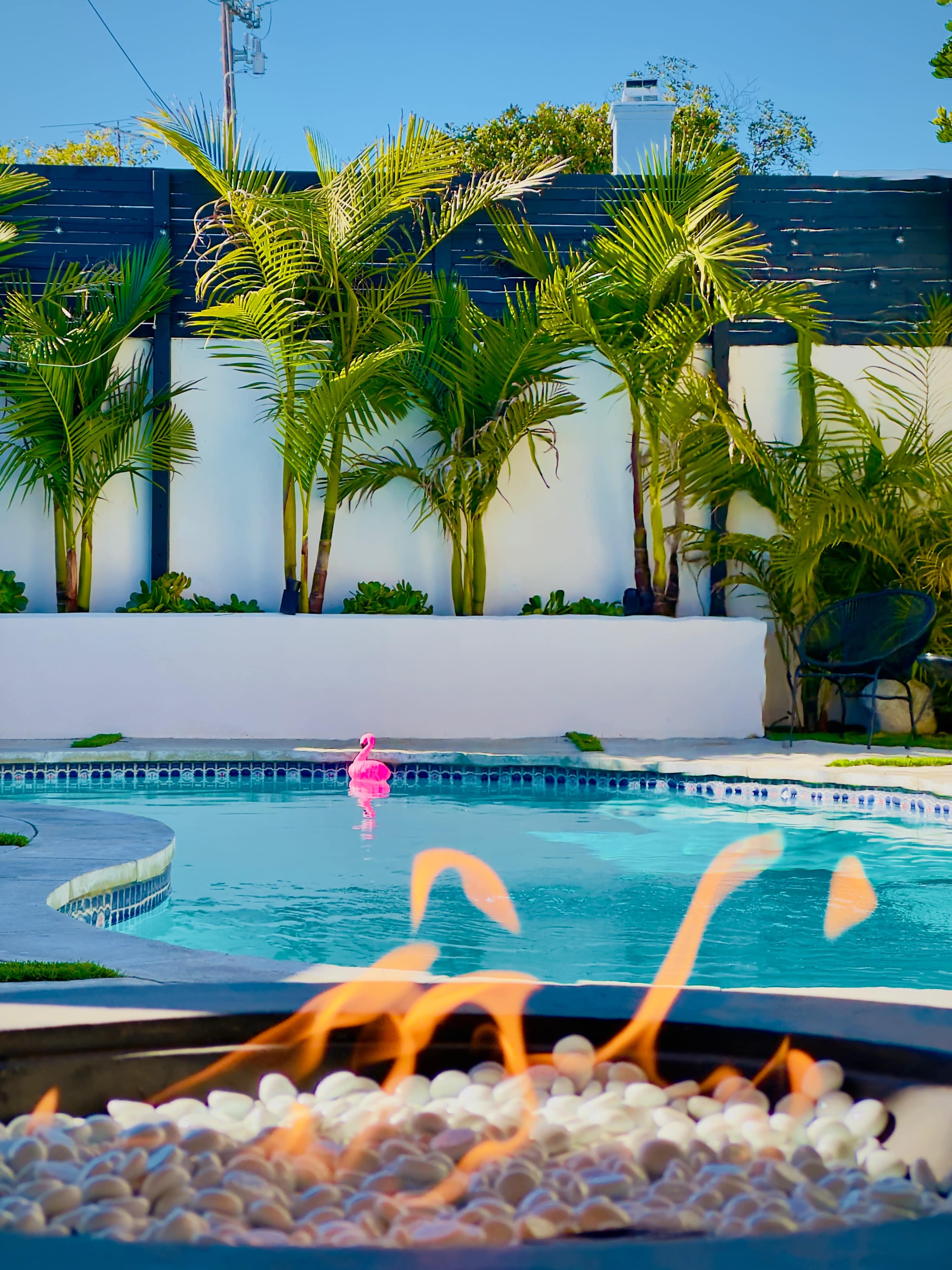 A pool with clear water surrounded by tropical plants and a pink inflatable flamingo, with a fire pit in the foreground.