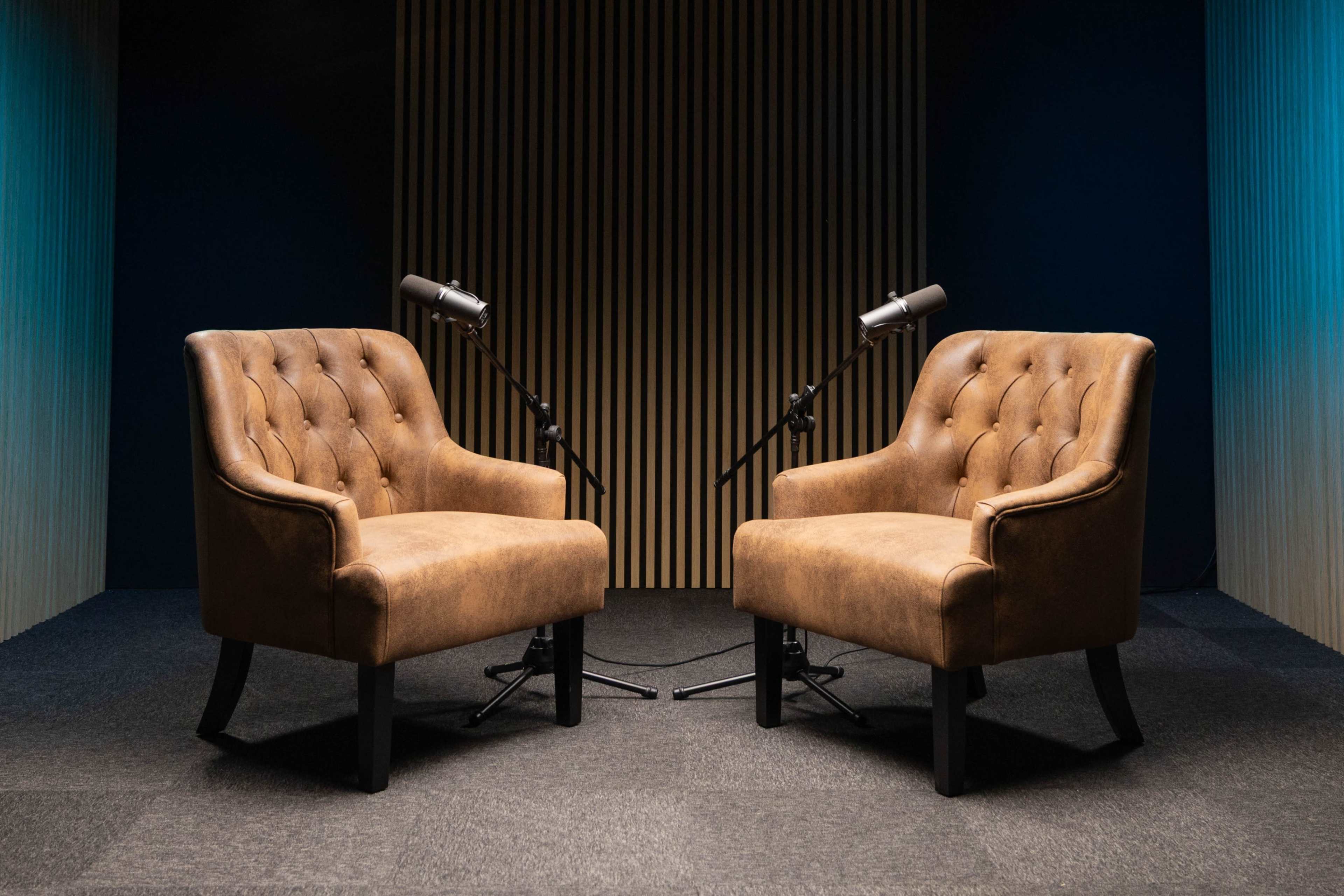 The image shows a pair of tufted brown armchairs facing each other with microphones positioned between them in a studio setting.