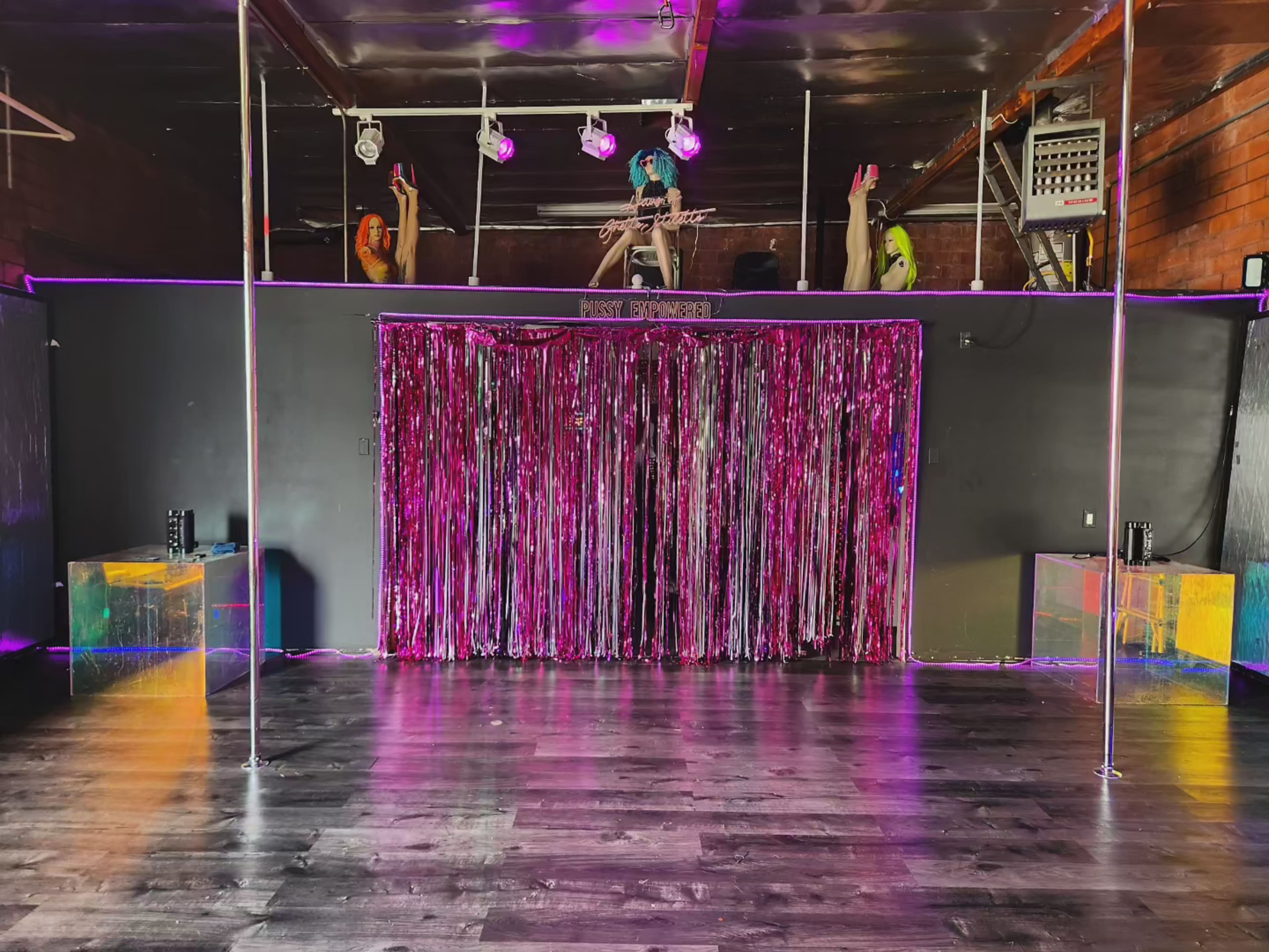 The image shows a dimly lit performance space with a polished wooden floor, featuring a shimmering pink curtain and illuminated poles against a dark backdrop.