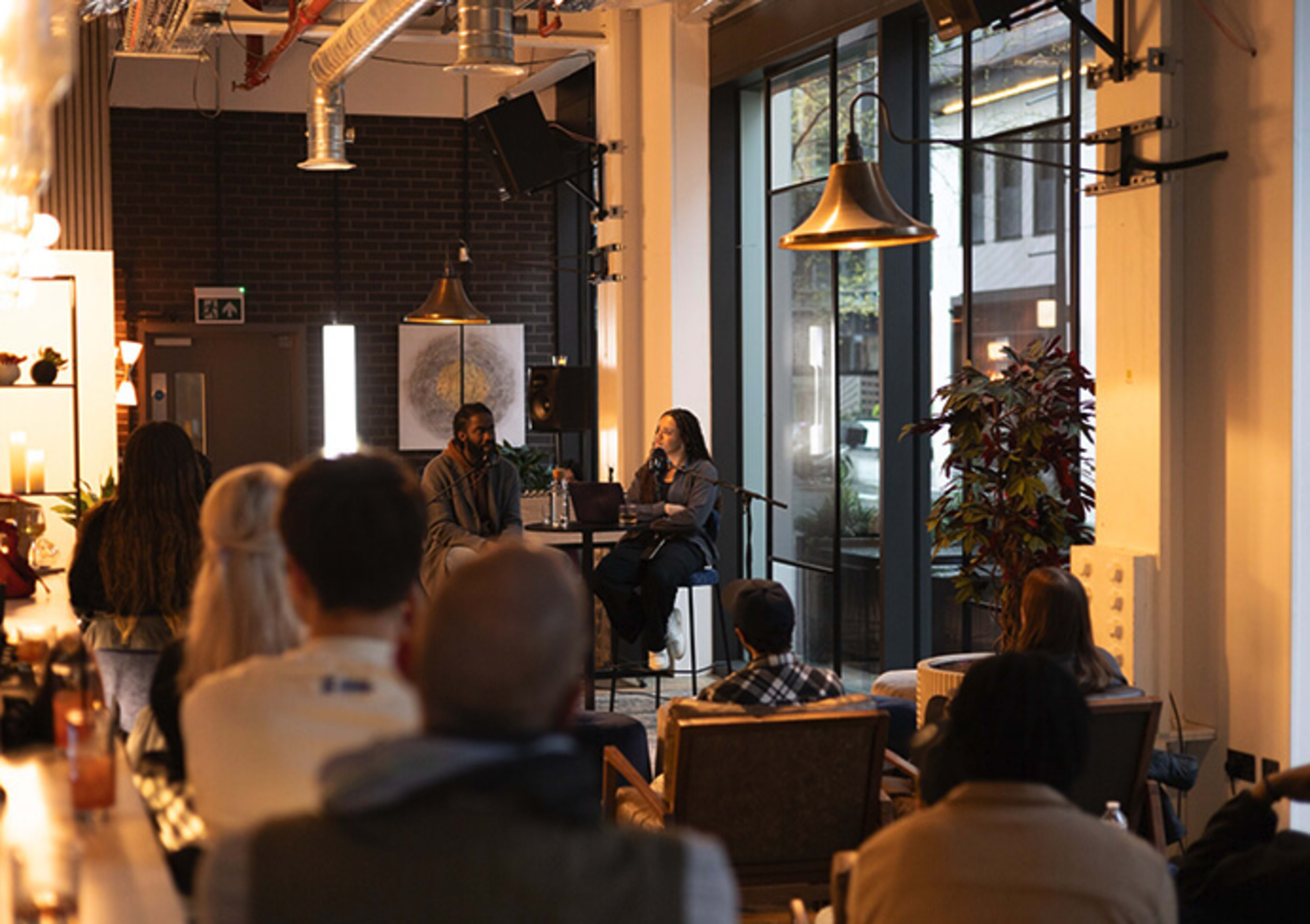 A panel discussion takes place in a modern indoor space with a seated audience listening to two speakers on stage.