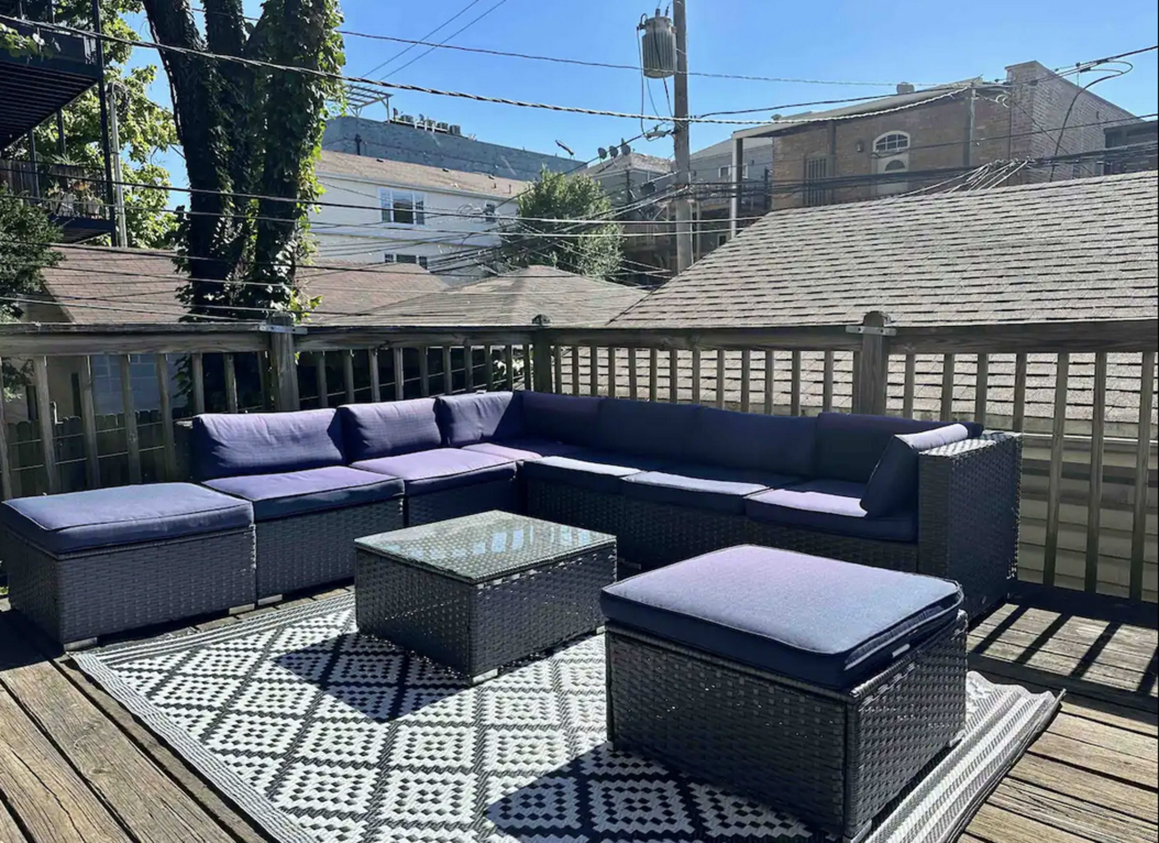 A patio deck featuring a large sectional sofa, a glass-top table, and a patterned rug in an outdoor setting with nearby houses in the background.