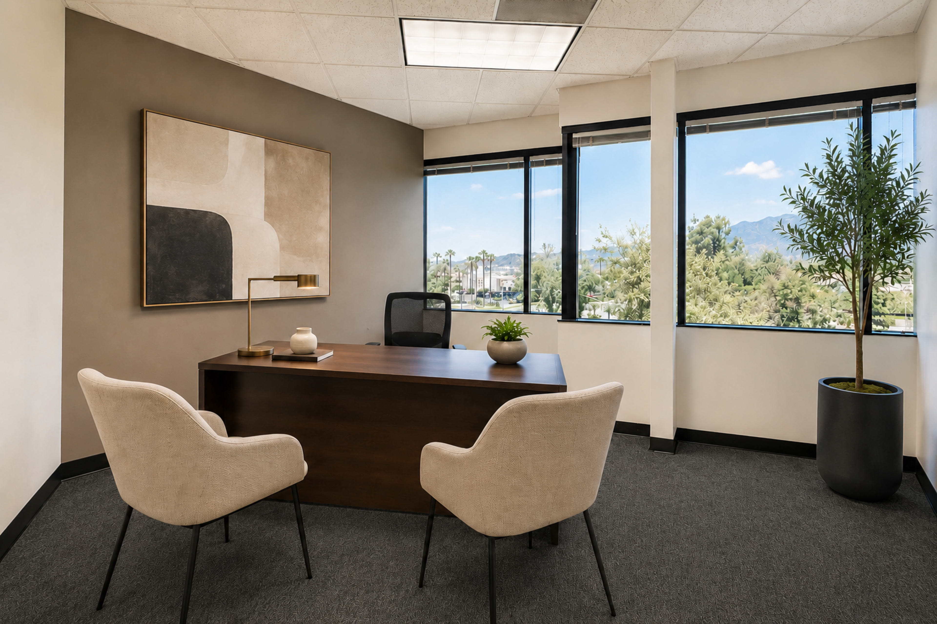 The image shows a modern office with a wooden desk, two upholstered chairs, and large windows offering a view of mountains and trees.