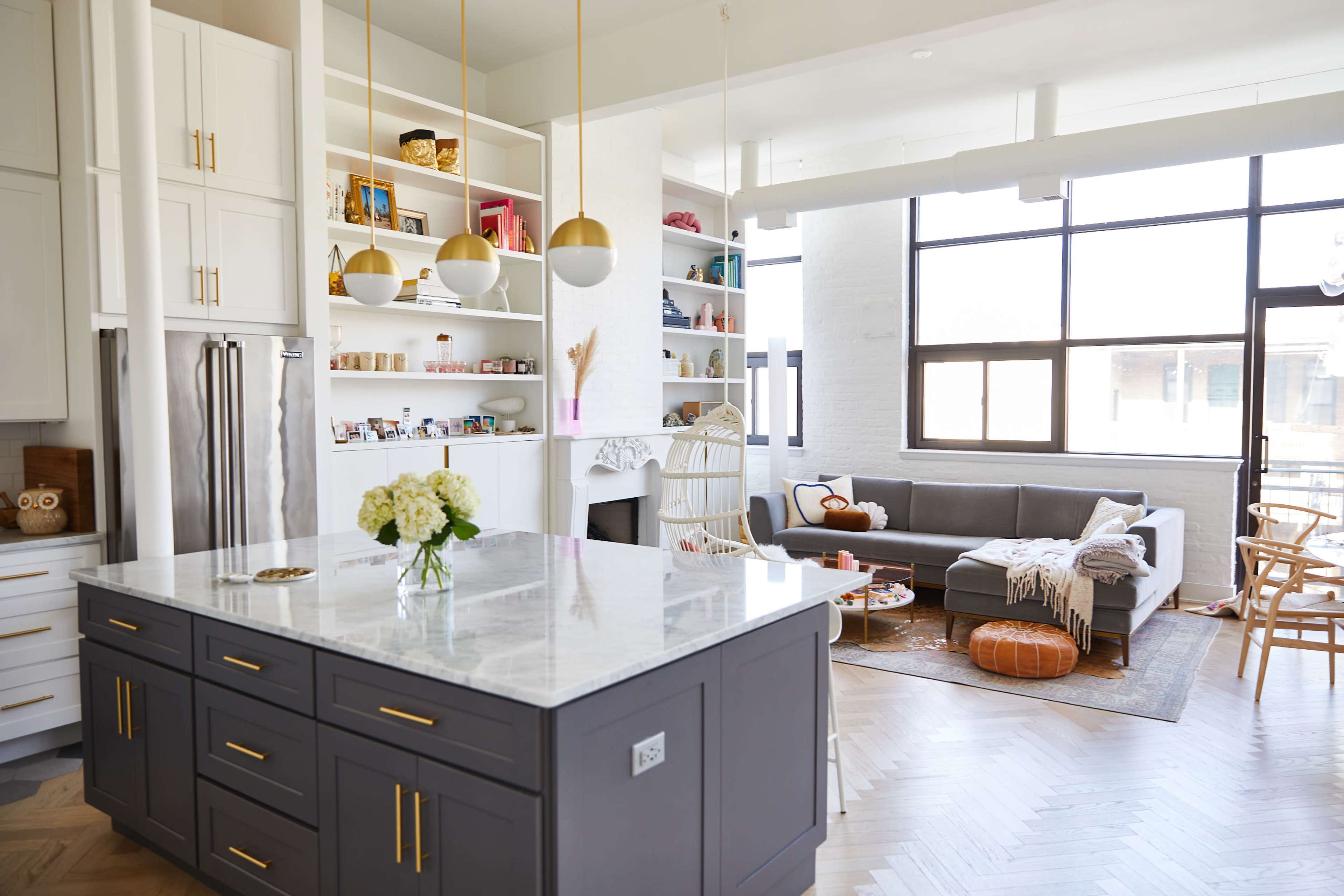 A modern kitchen features a large island with a marble countertop, surrounded by white cabinetry and open shelves, while a cozy seating area with a gray sofa is visible in the background.