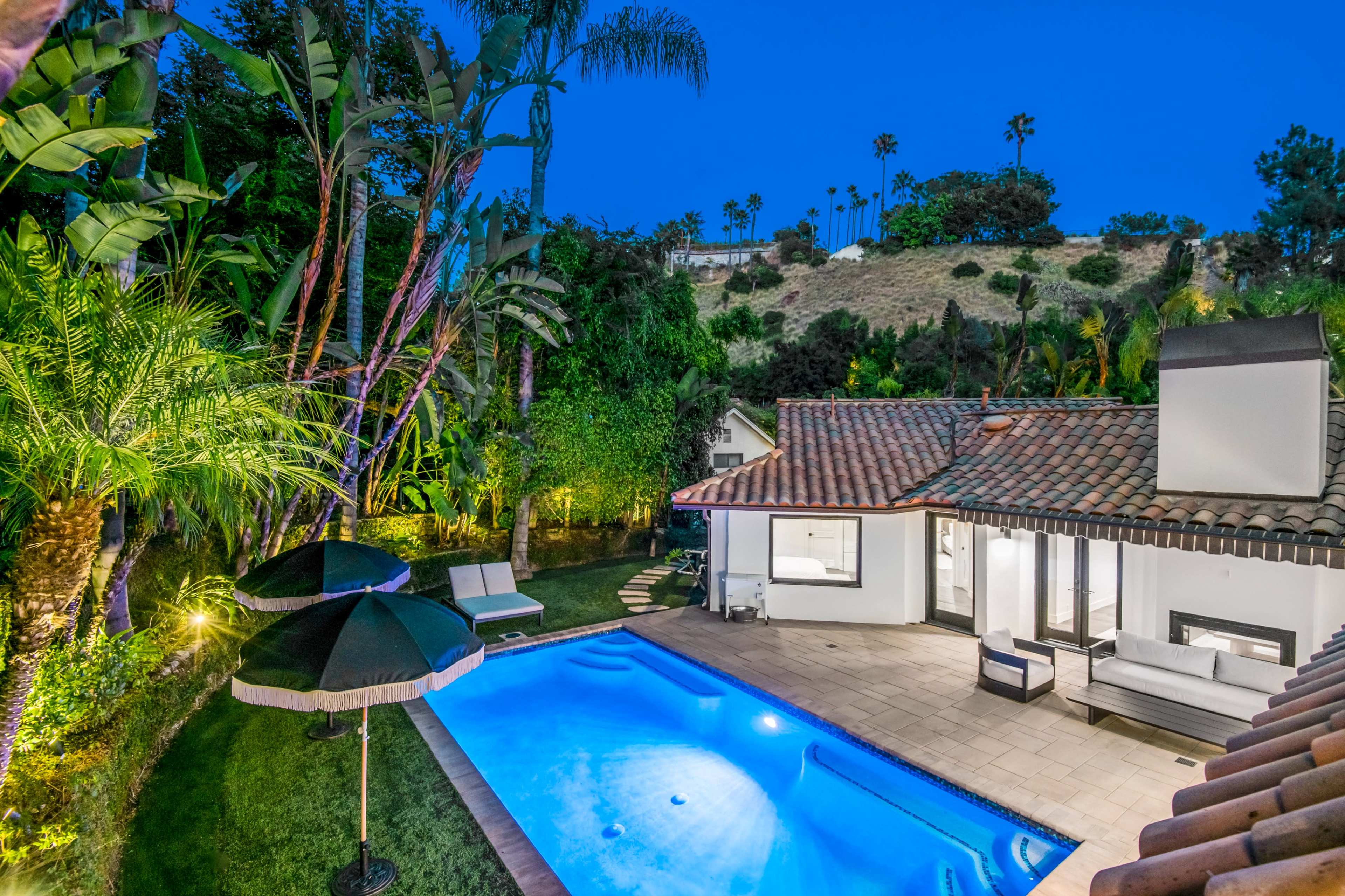 A landscaped backyard with a swimming pool, patio furniture, and tropical plants surrounded by palm trees at dusk.