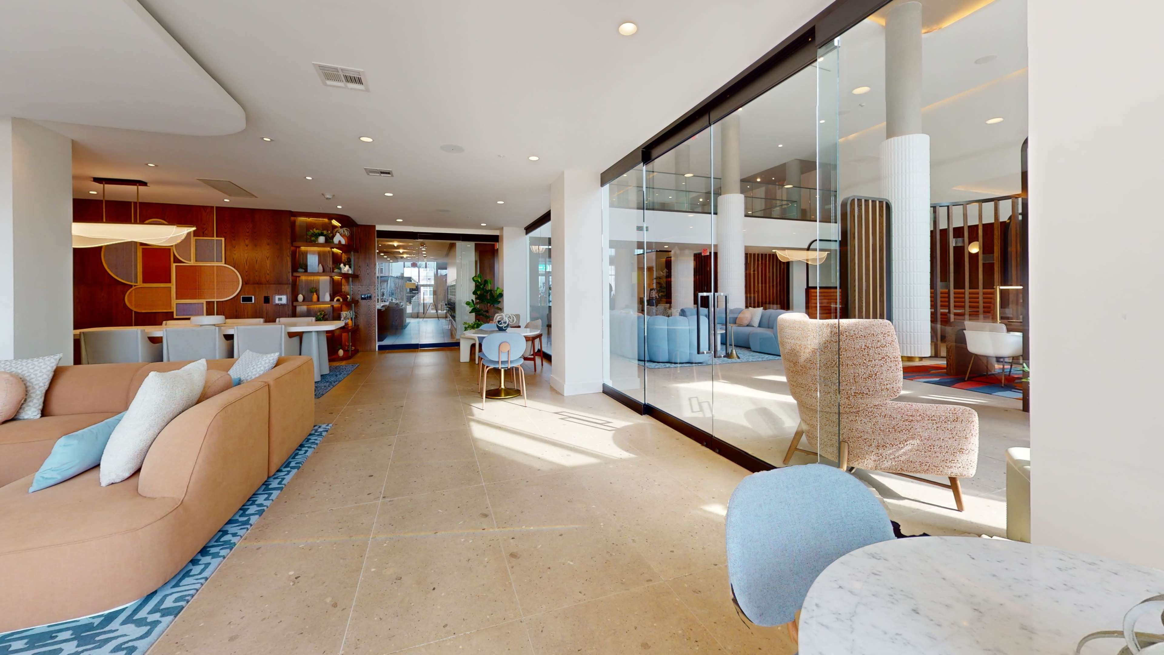 The image shows a modern interior space featuring a blend of seating areas, bookshelves, and large glass windows that allow natural light to enter.