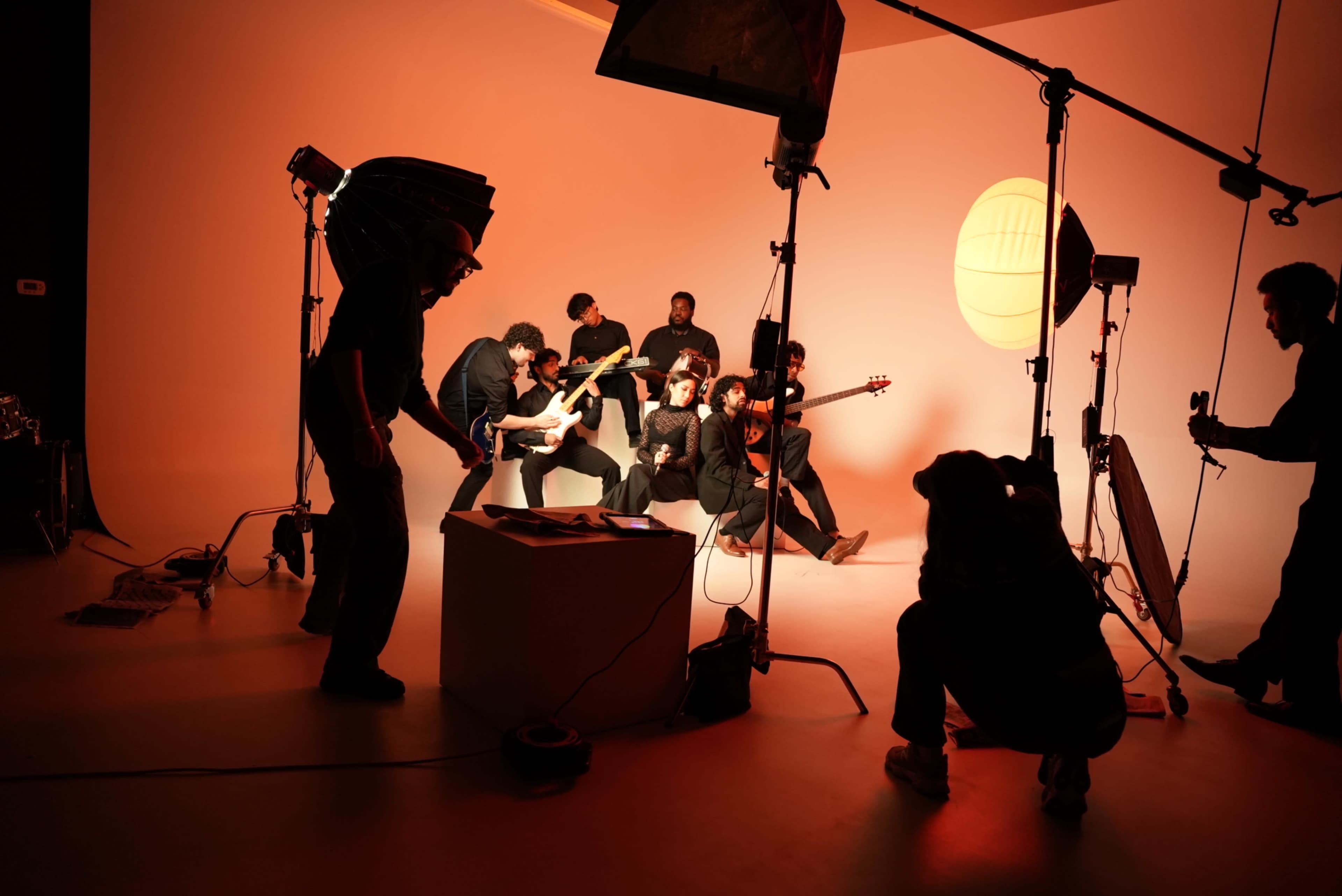 A group of musicians poses for a photoshoot in a studio with a backdrop featuring warm lighting and various photography equipment.