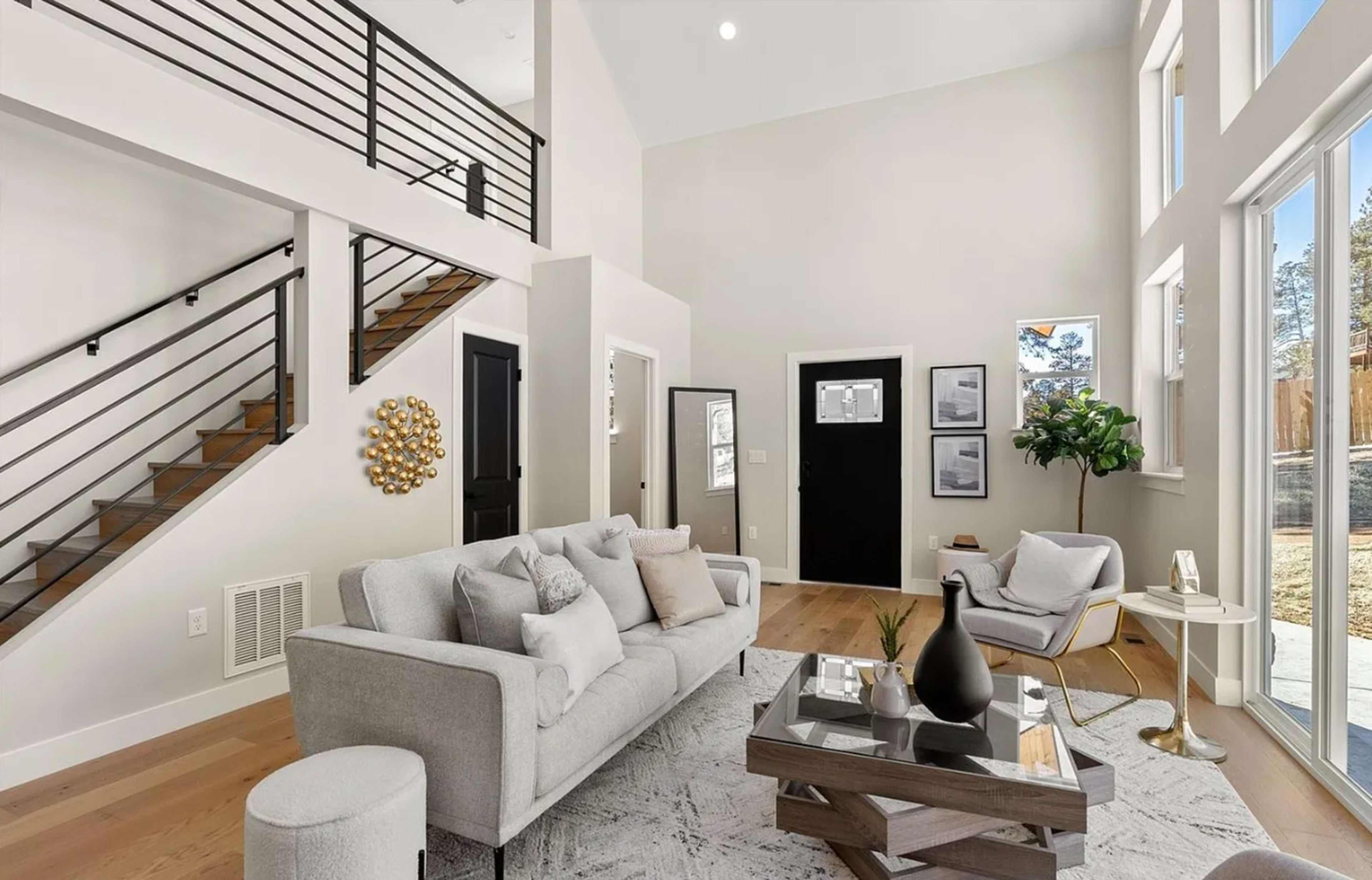 The image shows a modern living room with large windows, a light gray sofa, a coffee table, and a staircase in the background.