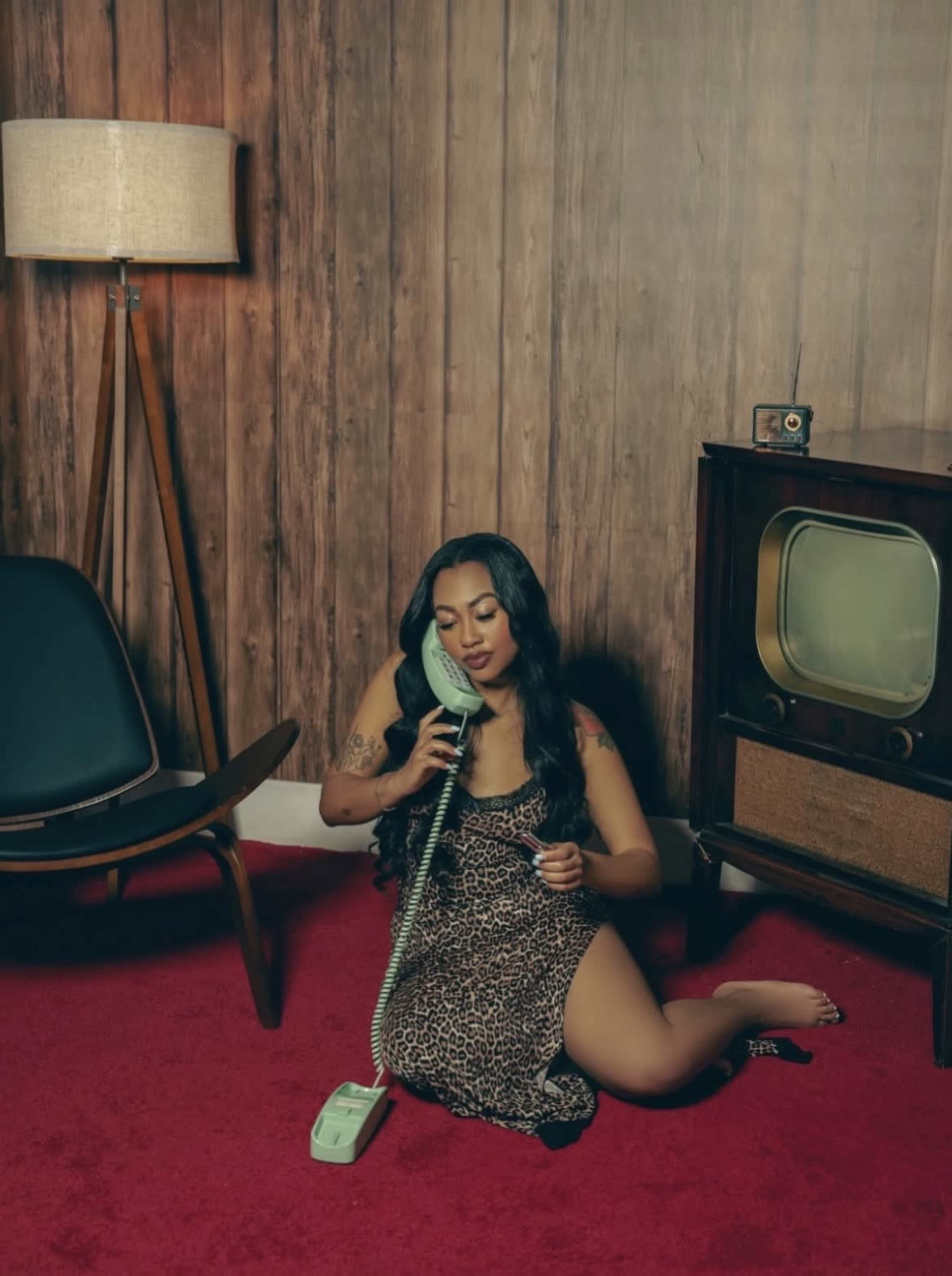 A woman in a leopard print dress sits on a red carpet while holding a green rotary phone, surrounded by mid-century furniture and a wooden wall.
