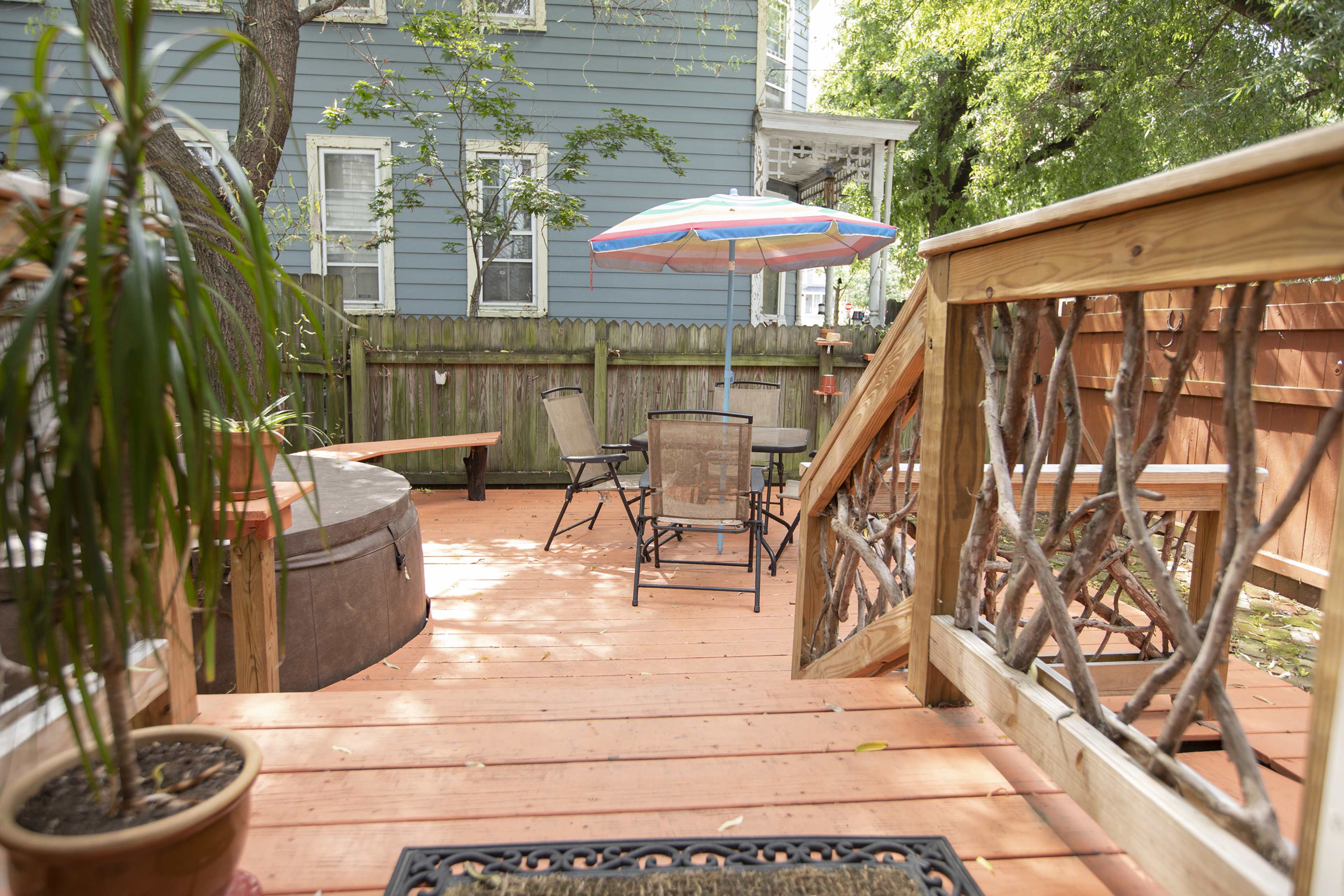 A wooden deck with a hot tub, a chair and table set, and a colorful umbrella, surrounded by greenery and a blue house in the background.