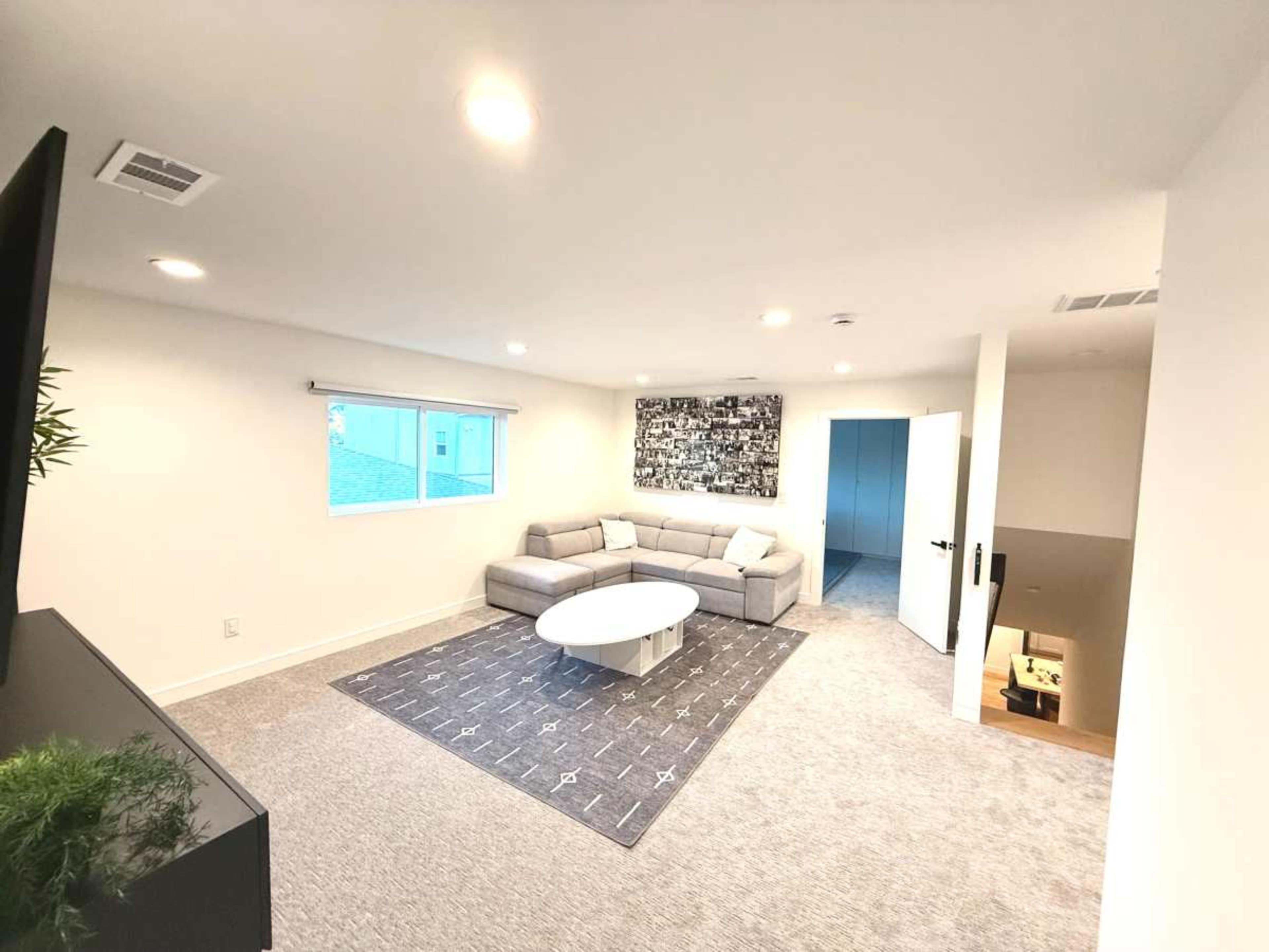 The image shows a modern living room with a gray sectional sofa, a round white coffee table, and a large wall art piece, with a window allowing natural light and a staircase in the background.