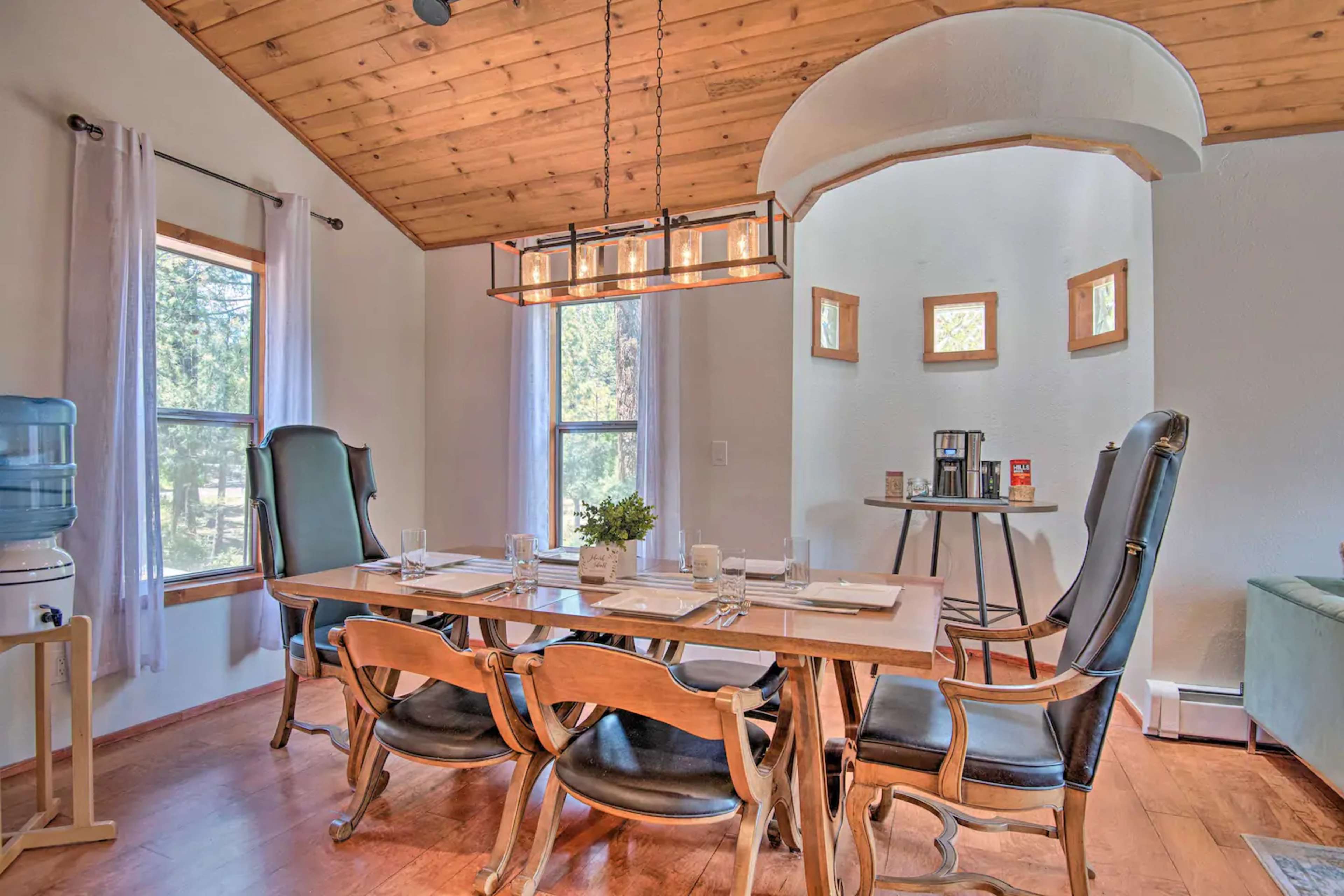 A dining area with a wooden table set for four, surrounded by chairs, under a beamed ceiling and illuminated by a rectangular light fixture.