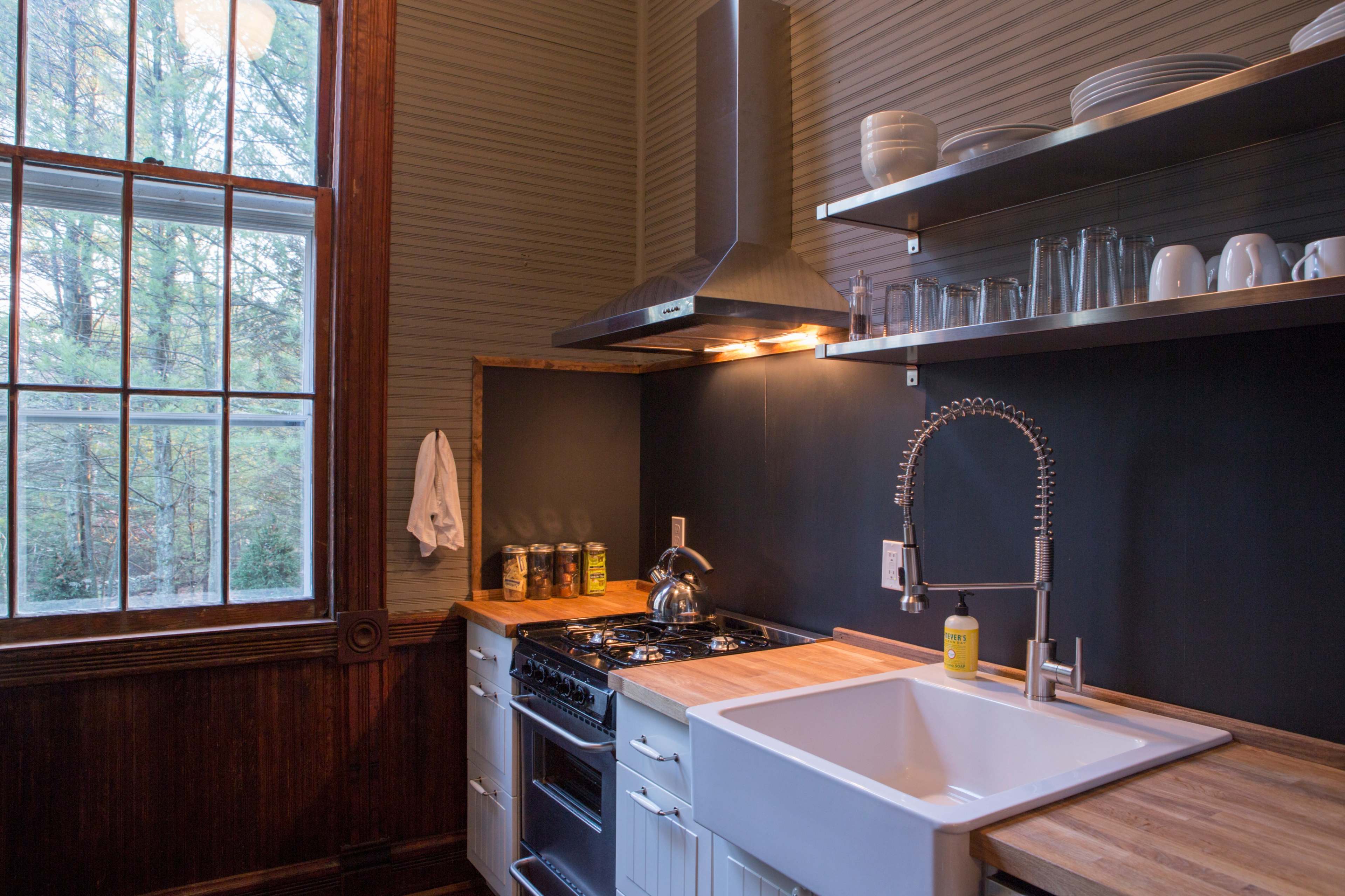 A contemporary kitchen features a large sink, a stovetop with a hood, and shelves displaying glassware and jars, with a large window providing natural light.