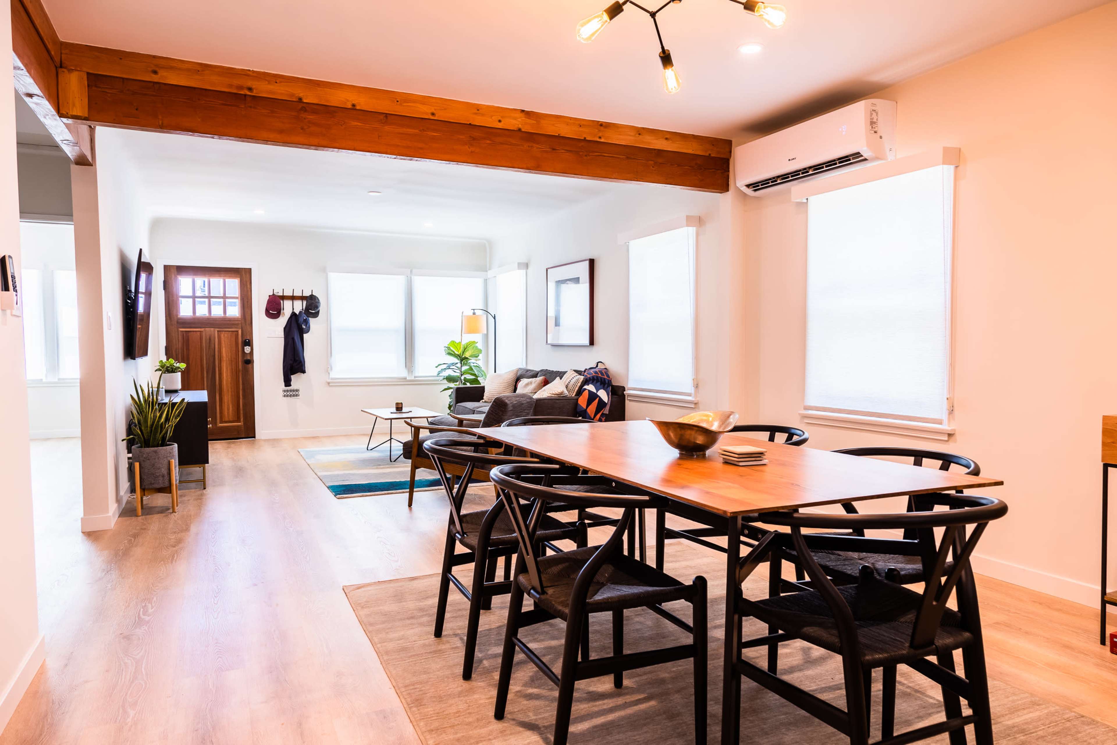 The image shows a modern, open-concept living area with a dining table, seating area, and wooden beams across the ceiling.