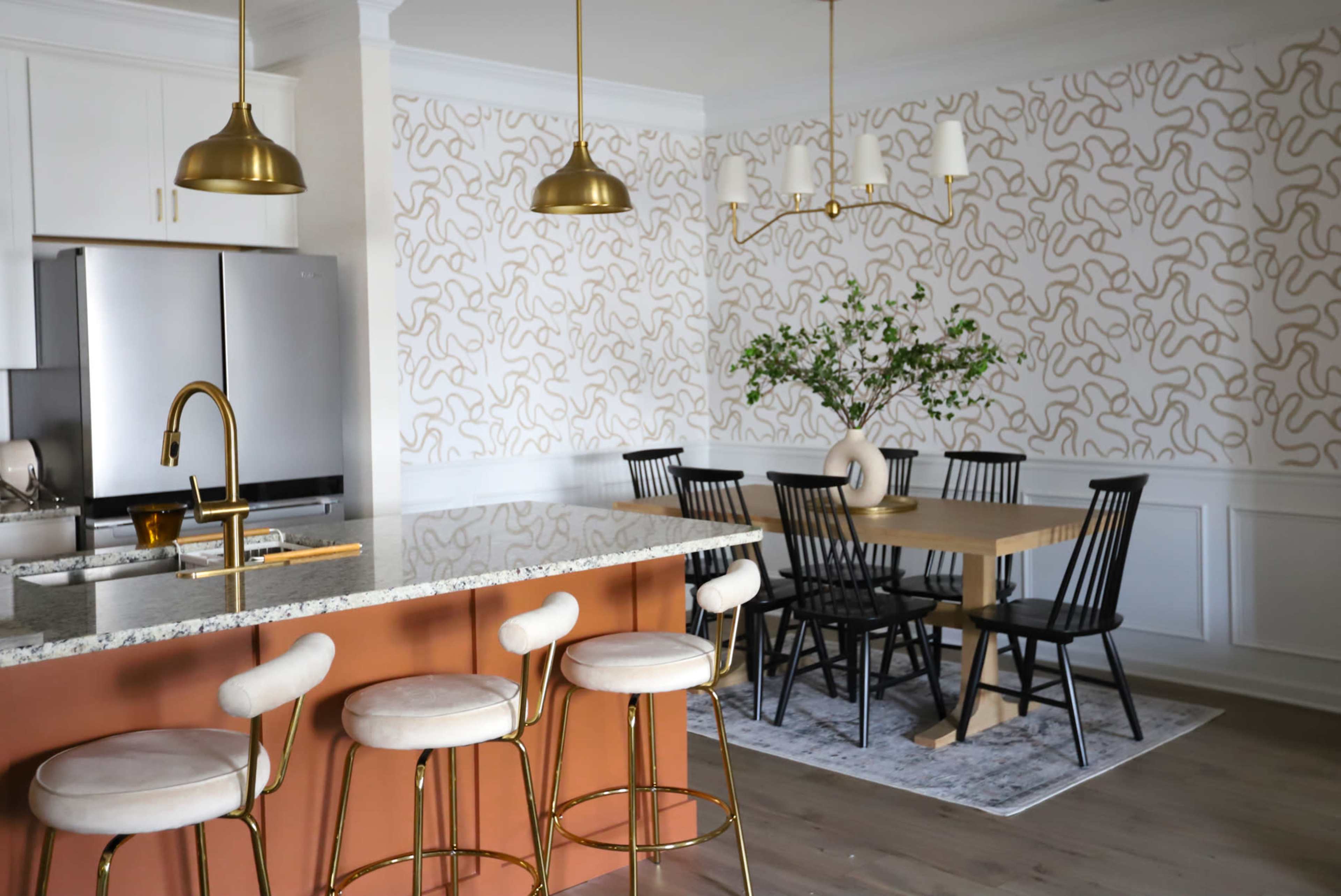 A modern kitchen and dining area feature a granite island with gold bar stools, a wooden dining table surrounded by black chairs, and patterned wallpaper.