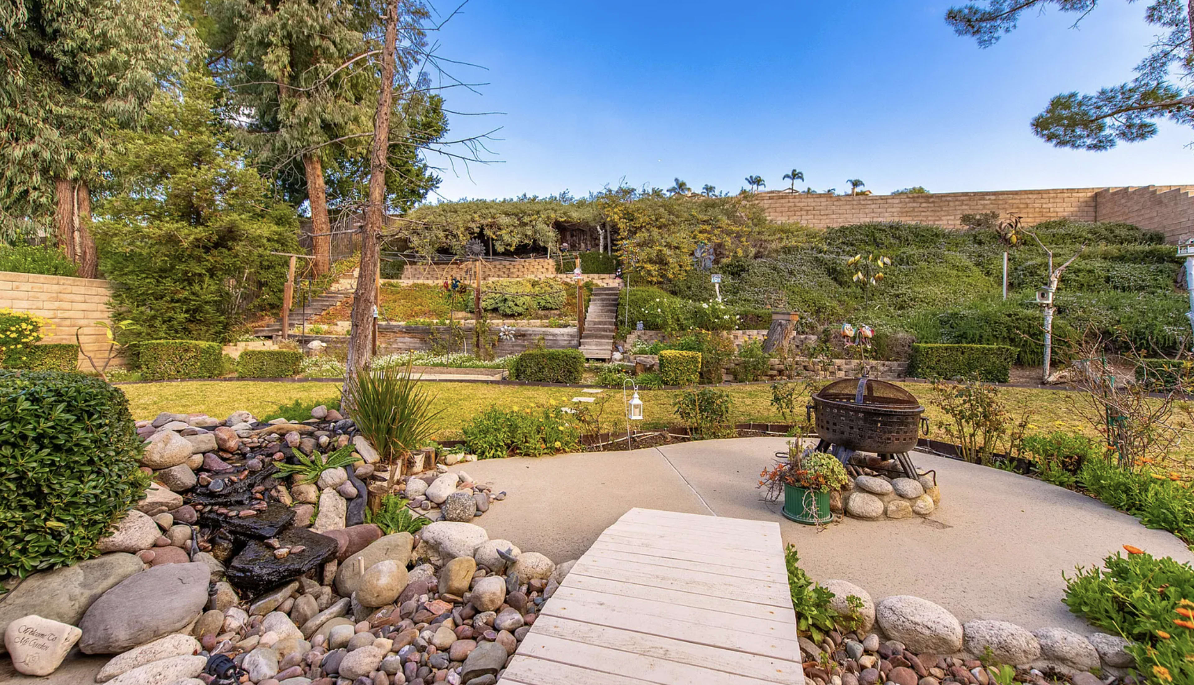 The image shows a landscaped garden with a stone pathway leading to terraced areas, greenery, and a fire pit.