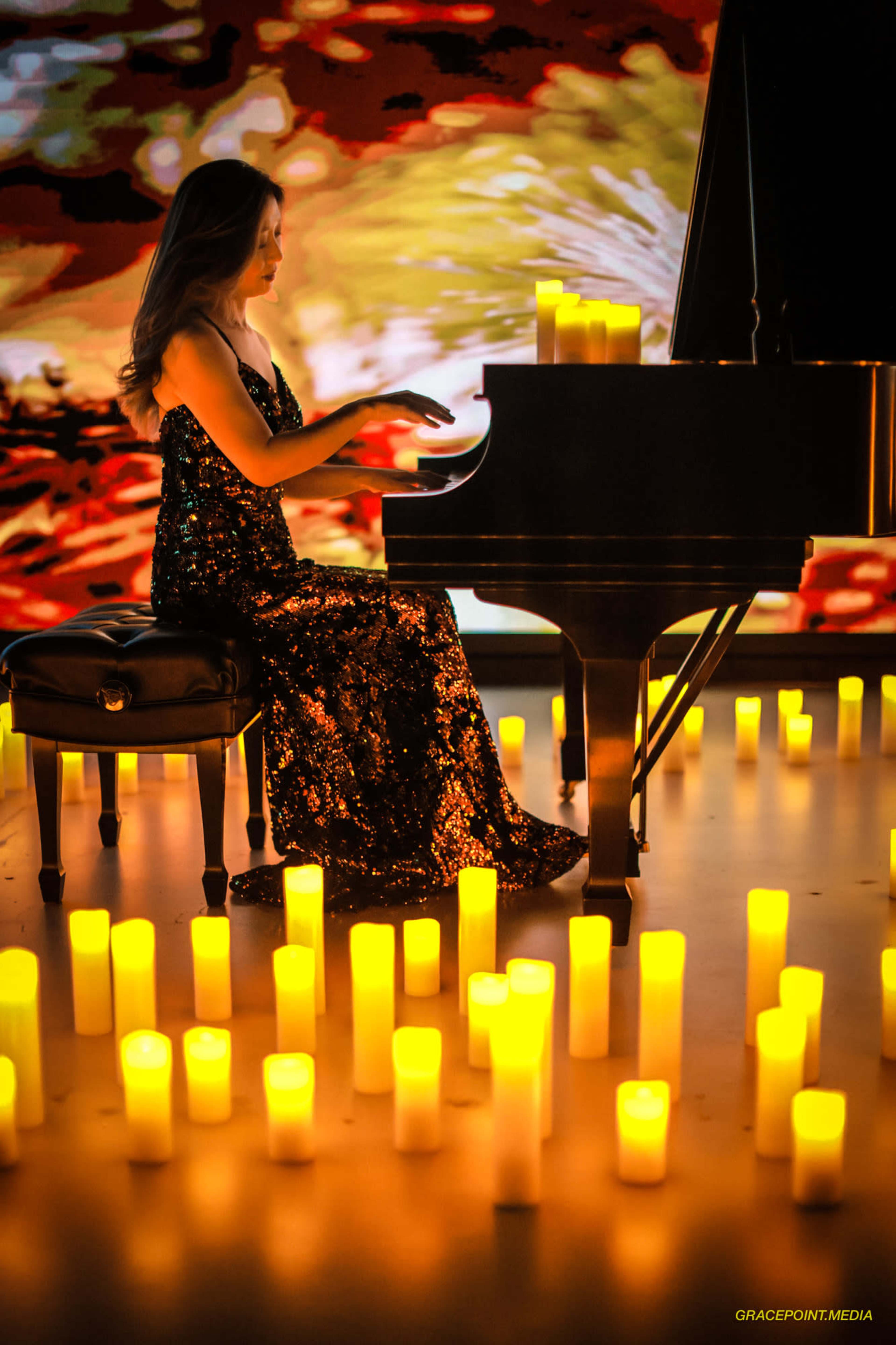 A woman in a sequined dress plays a piano surrounded by glowing candles on the floor.