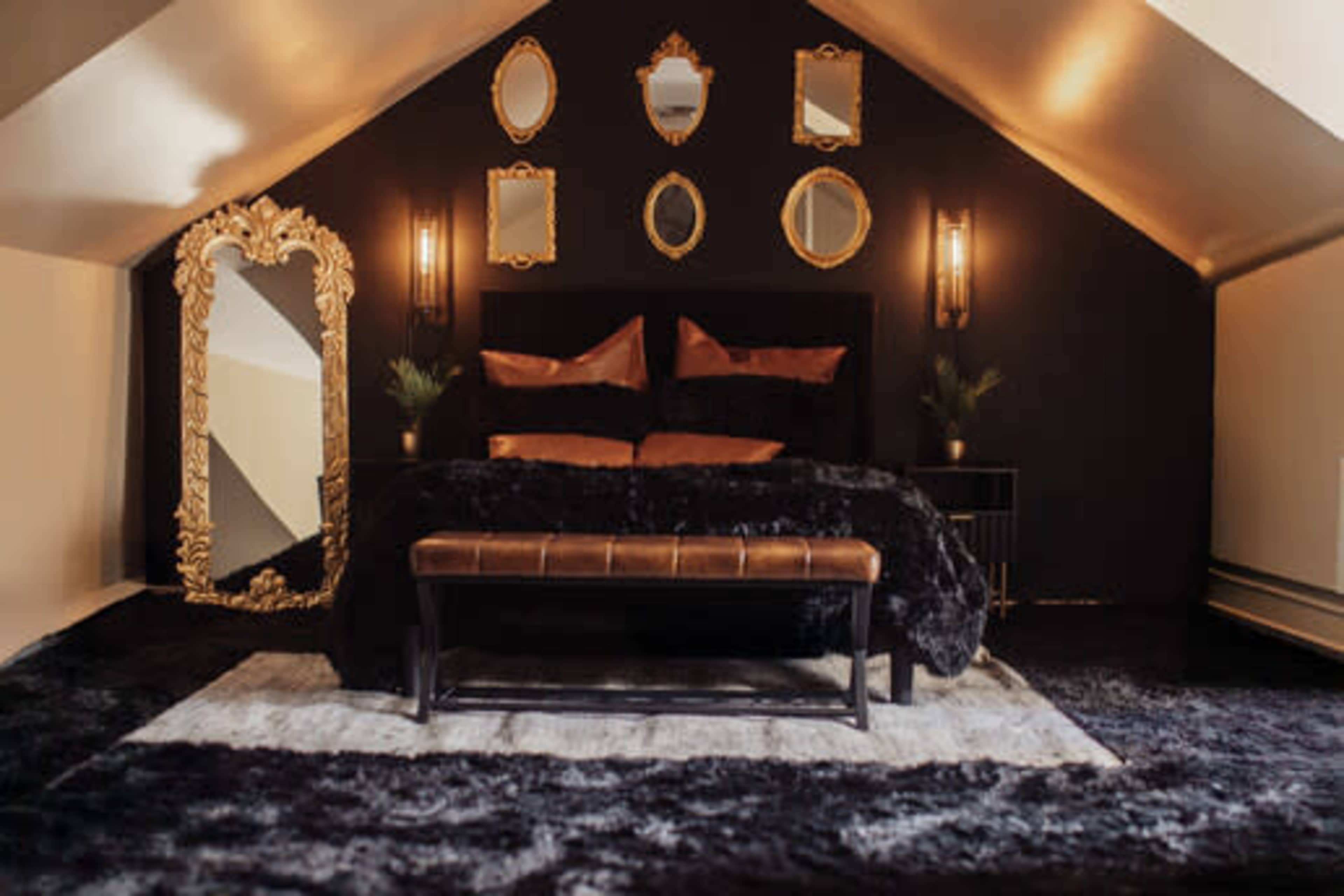 The image depicts a stylish bedroom with a black accent wall, a large mirror, and multiple decorative mirrors above the bed, which is dressed in dark bedding and accompanied by a bench at the foot.