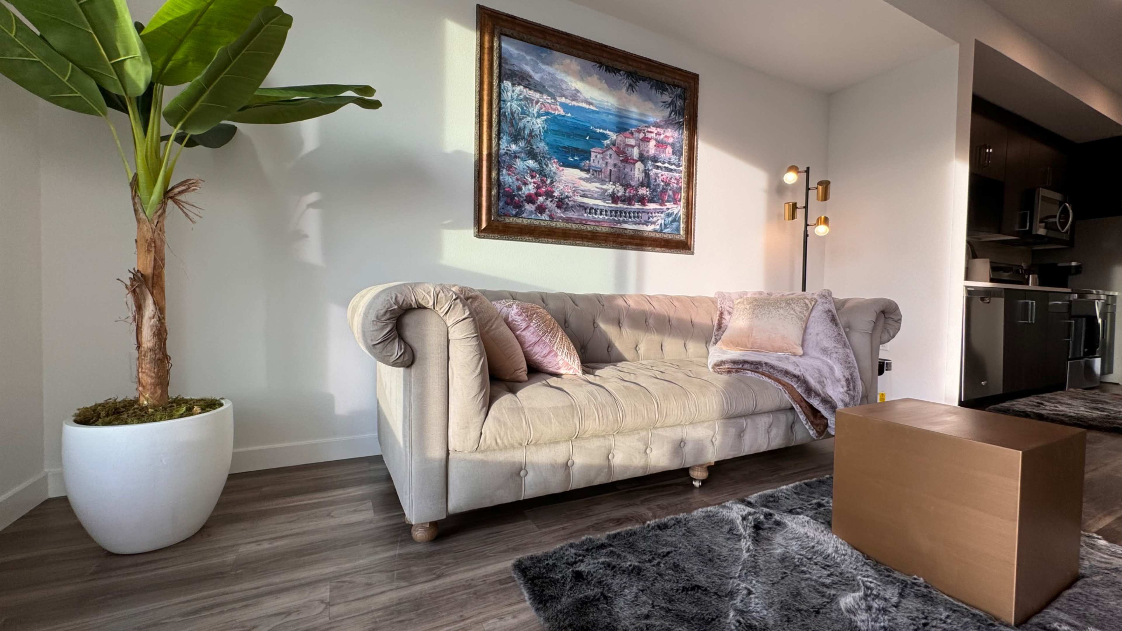 A beige tufted sofa with a decorative pillow and a cozy throw blanket, positioned near a potted plant and a gold-colored coffee table on a gray rug, with a framed painting above the sofa.