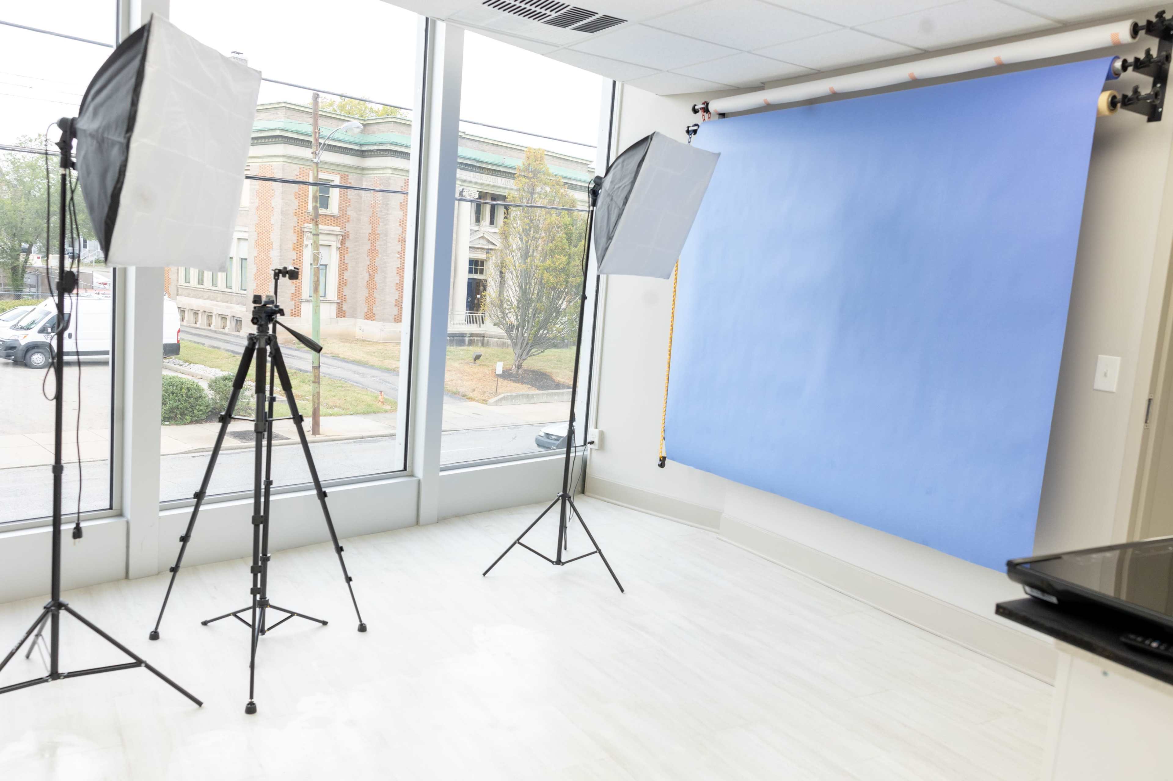 A photography studio features two softbox lights and a blue backdrop, with a camera tripod set up in a well-lit room with large windows.