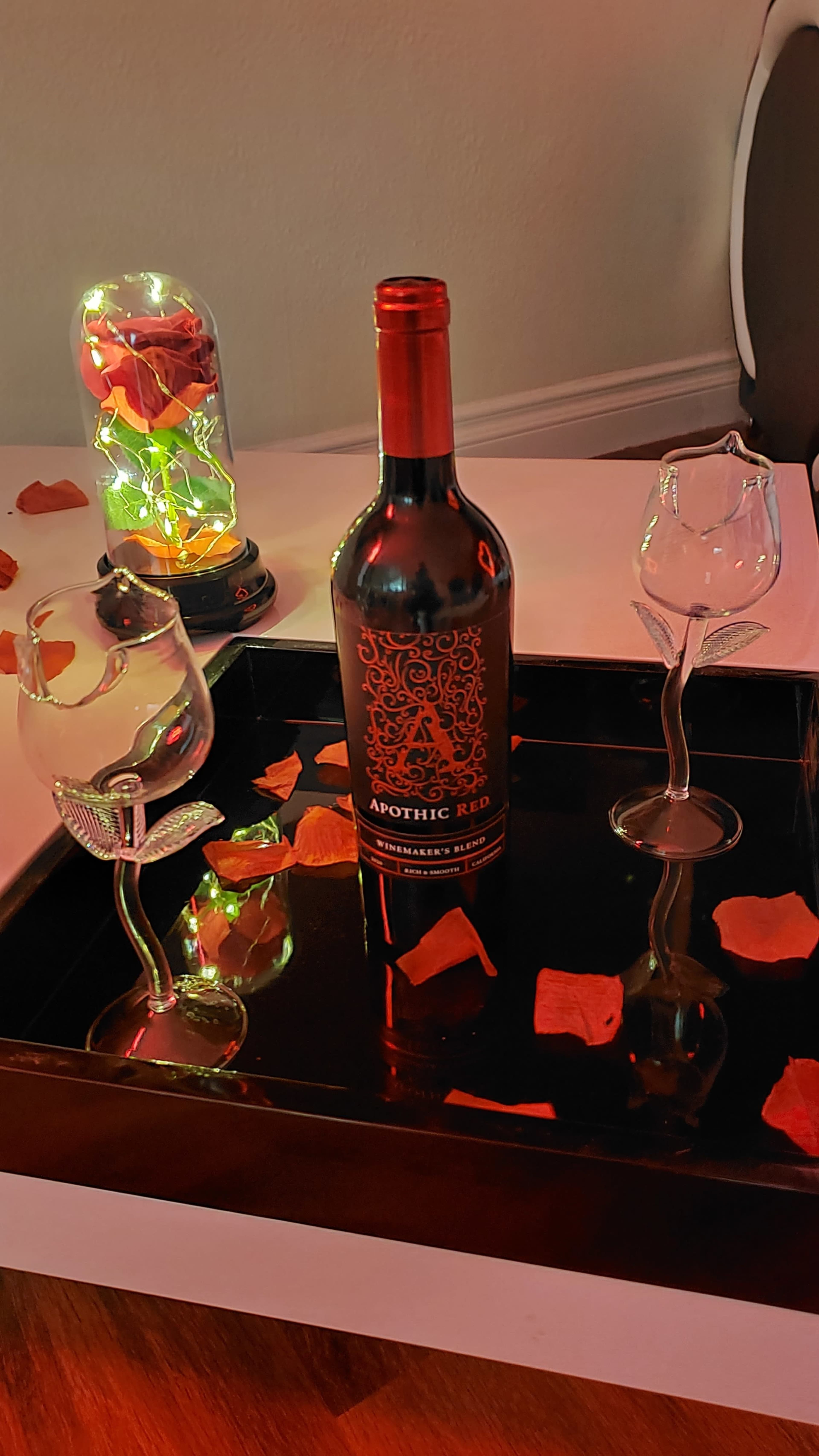A bottle of Apothic Red wine sits on a tray adorned with rose petals and two decorative glasses.