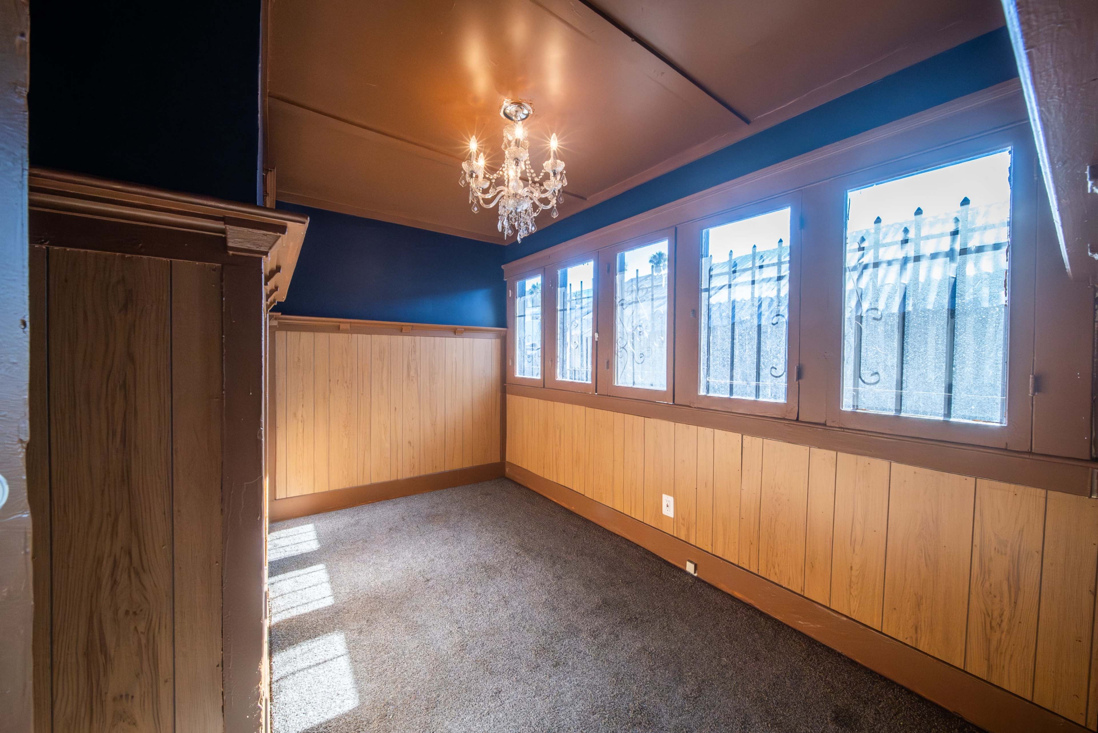 A small room features wooden paneling, a chandelier, and large windows with a view of a fence outside.