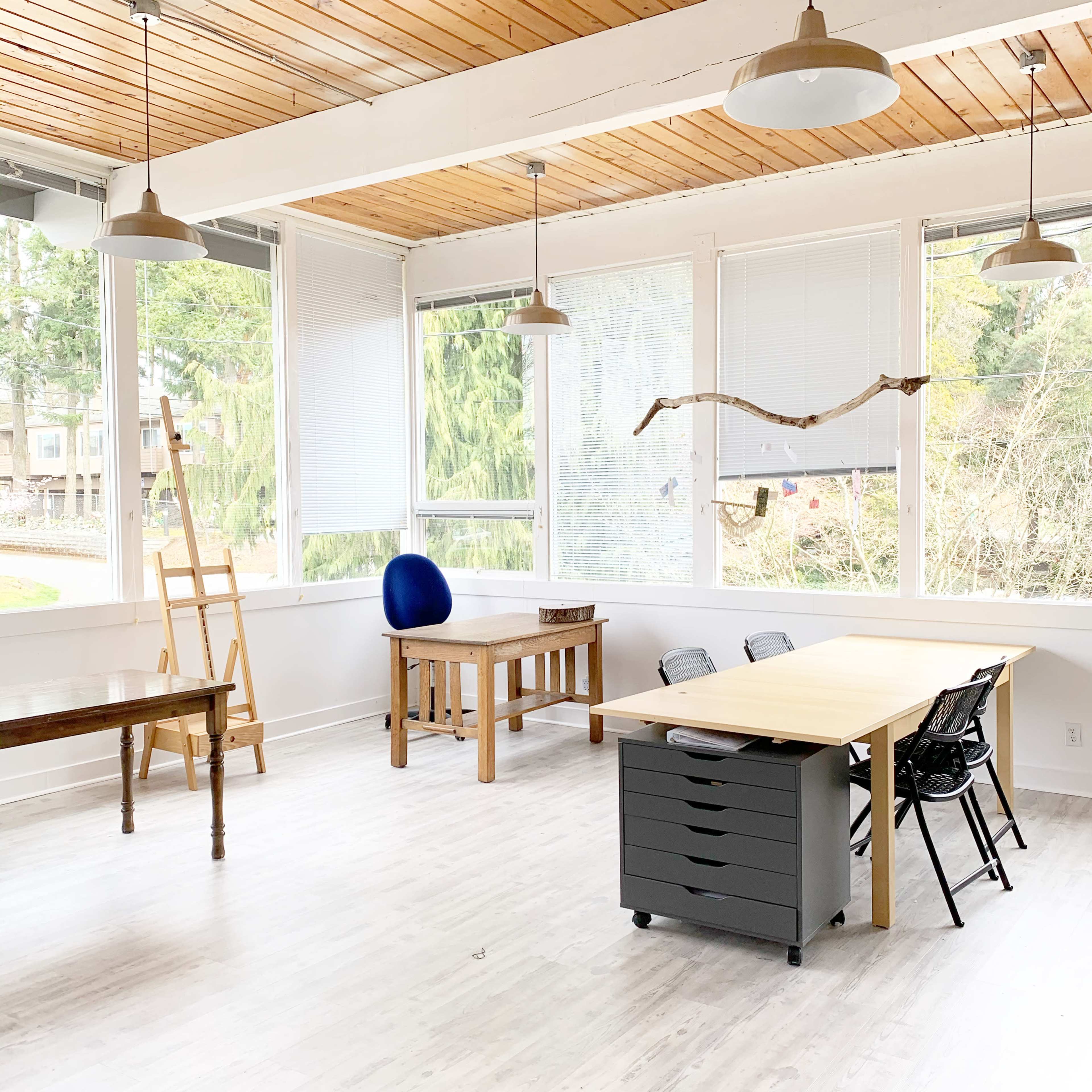 A bright room with large windows, featuring several wooden tables, a blue chair, and a storage unit with wheels.