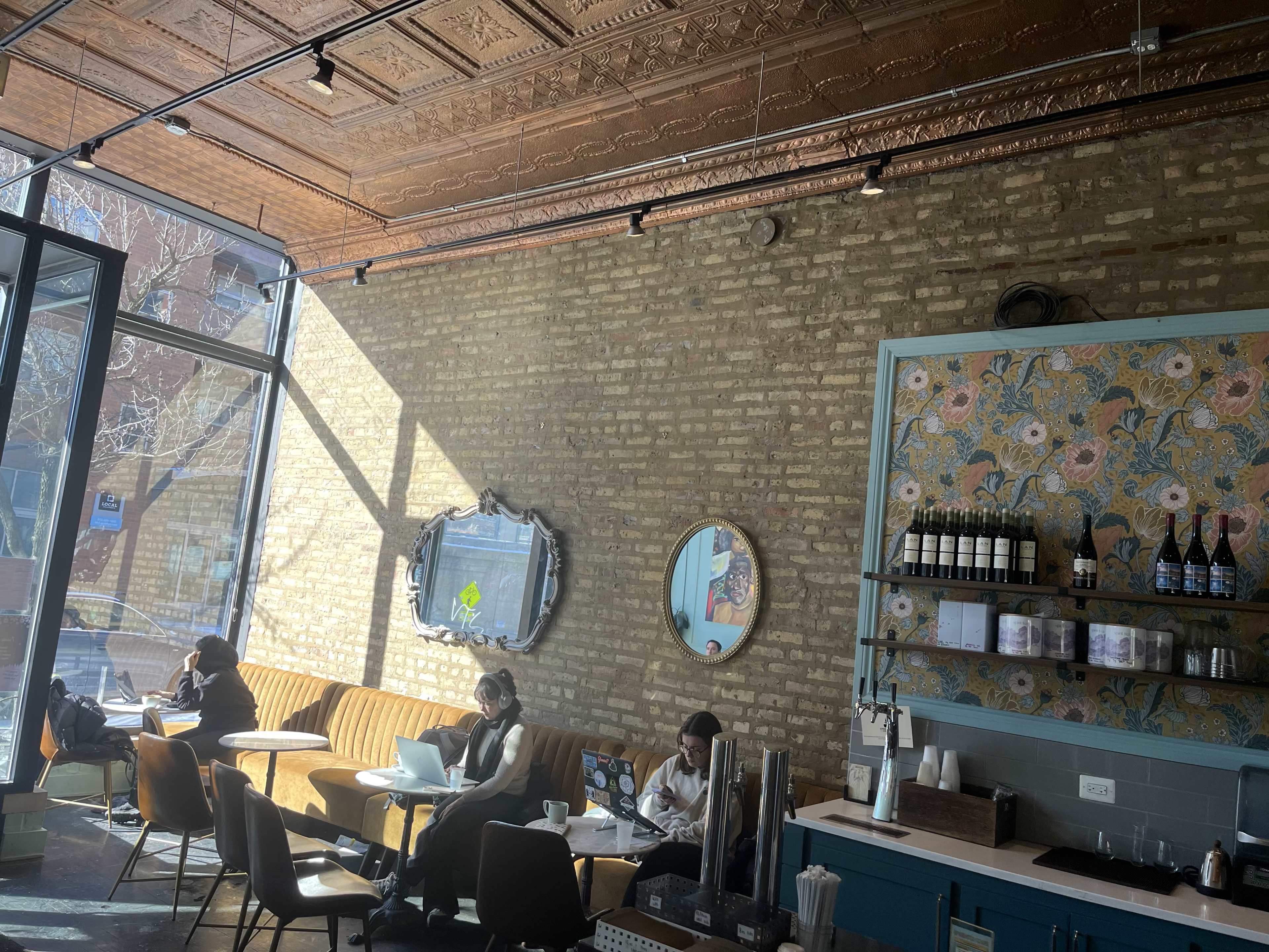 The image shows a cozy café with a brick wall, large windows allowing natural light in, and several patrons sitting at tables working on laptops.