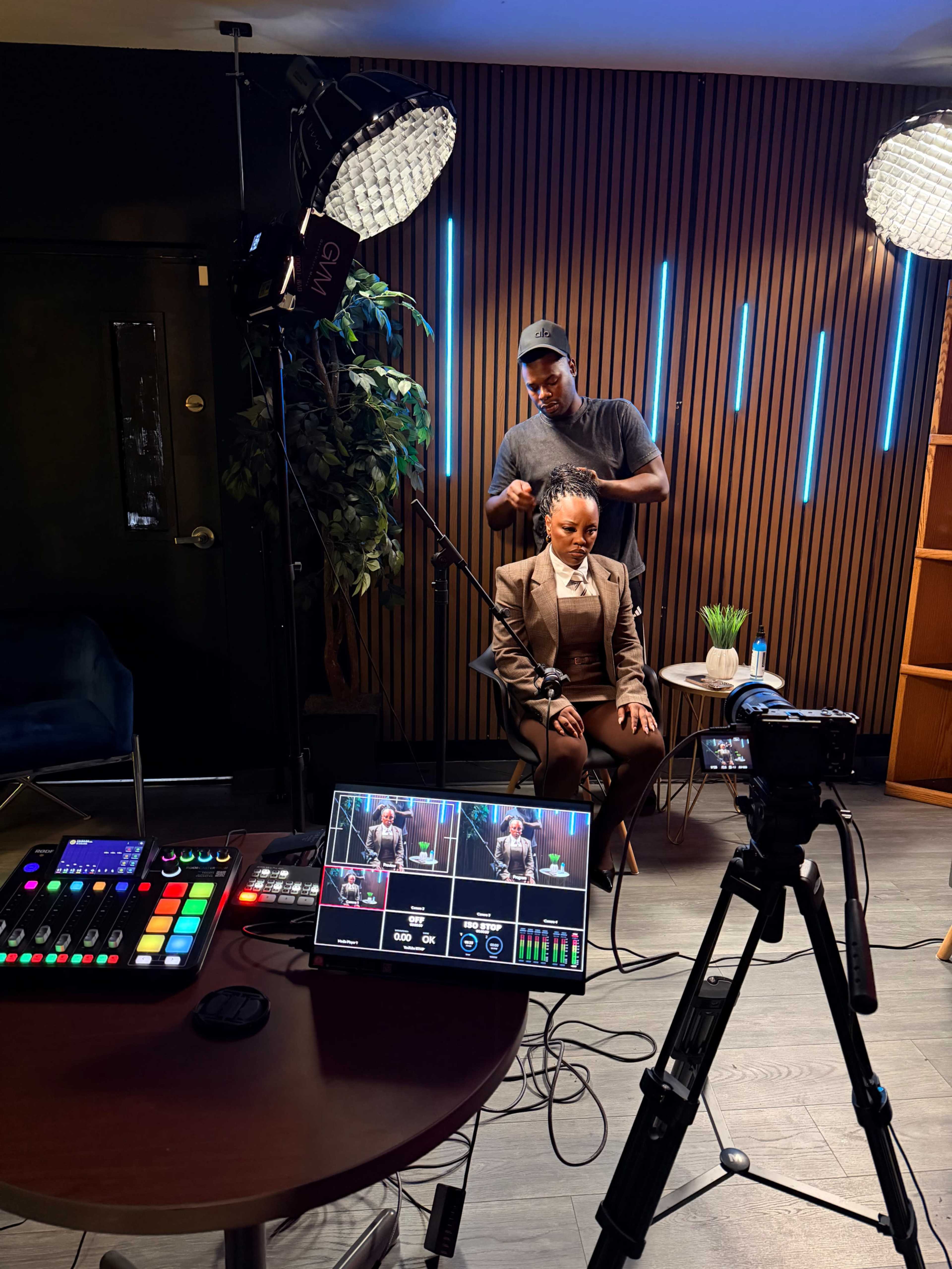 A person is seated in front of a camera setup while a stylist adjusts their hair in a modern studio with wooden panels and lighting equipment.