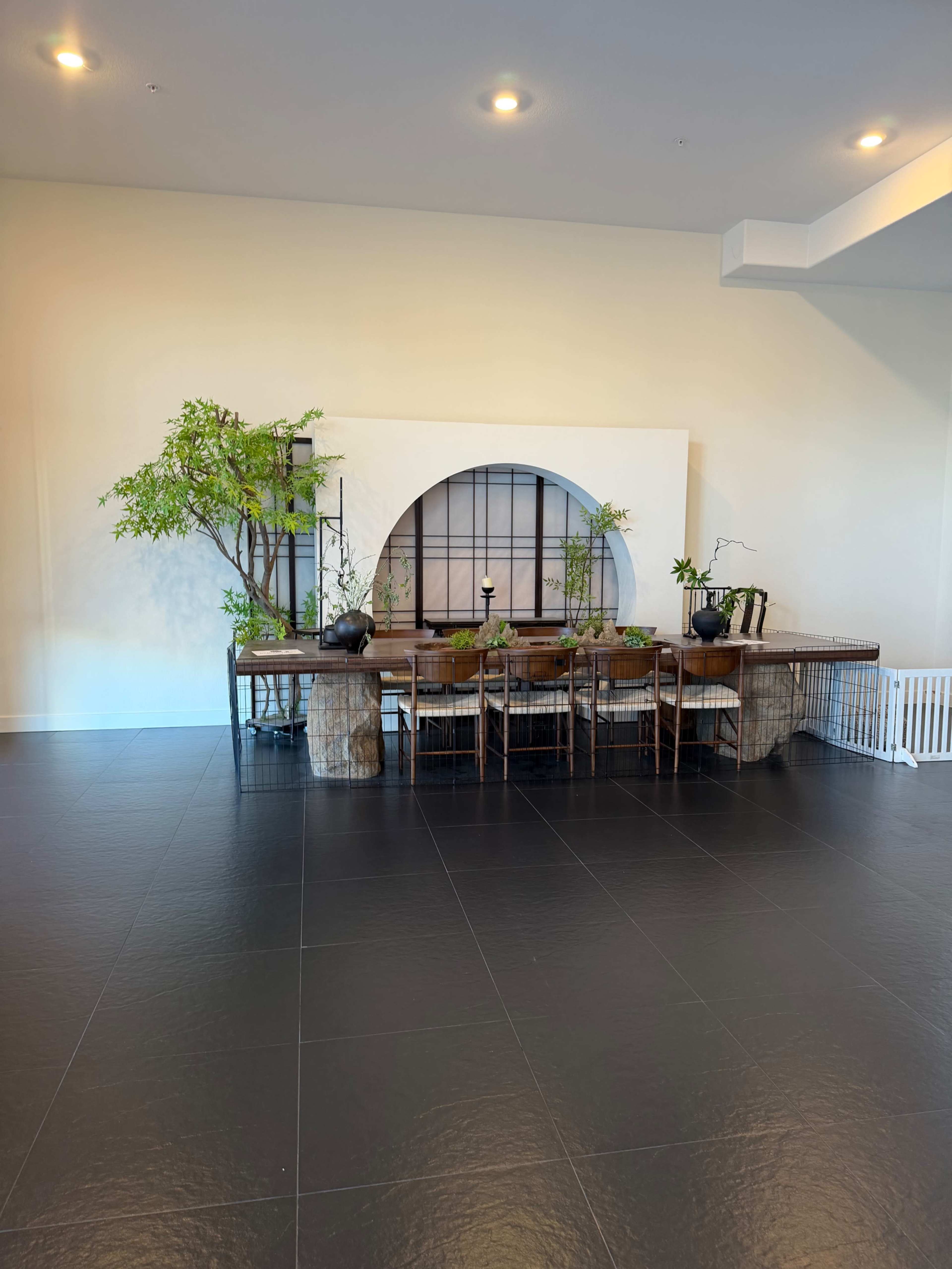 The image shows a spacious room with a wooden table adorned with potted plants, positioned against a backdrop featuring a large circular design and additional greenery.