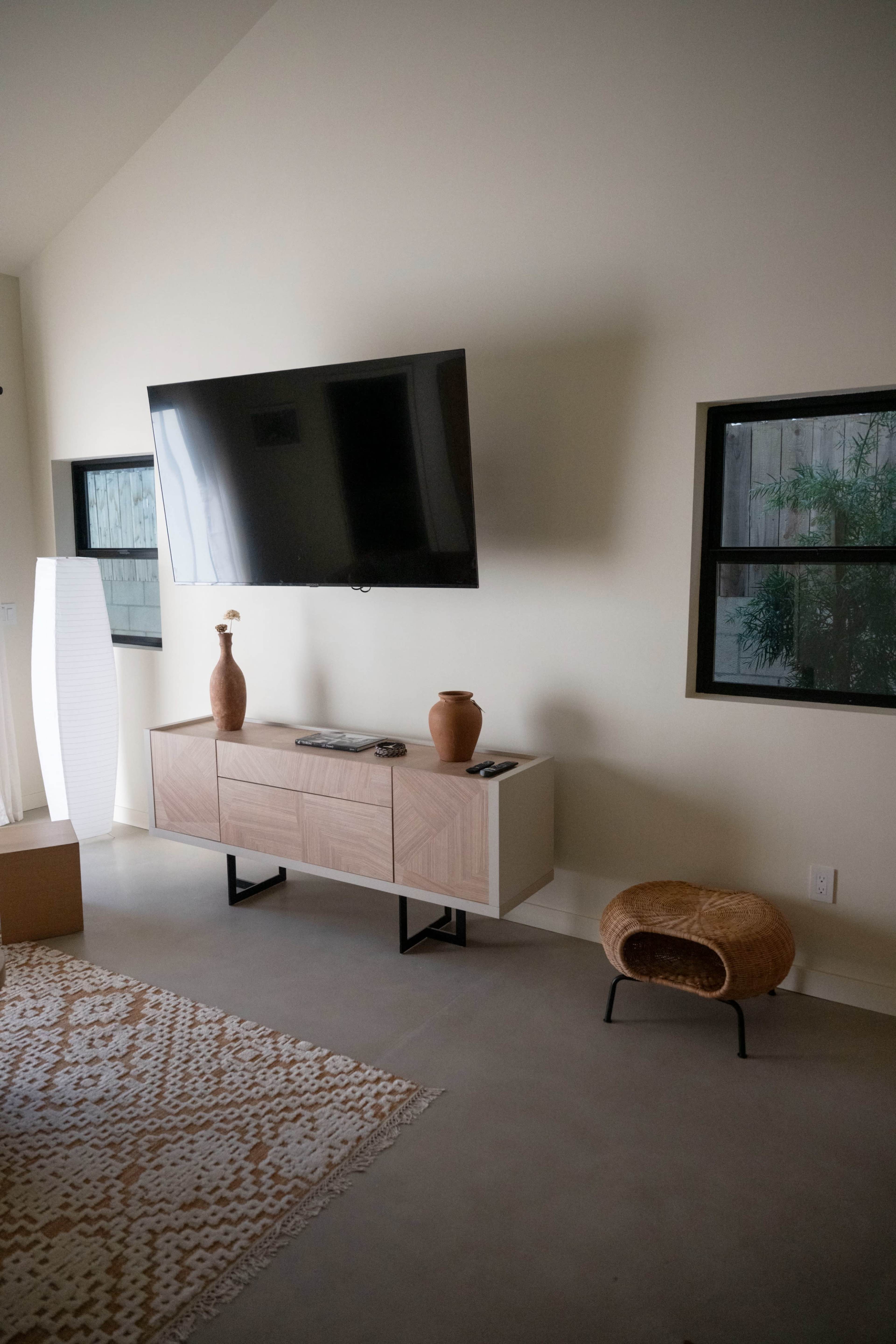 The image shows a modern living room with a mounted television above a wooden cabinet, a decorative vase, and a small woven stool beside a window.