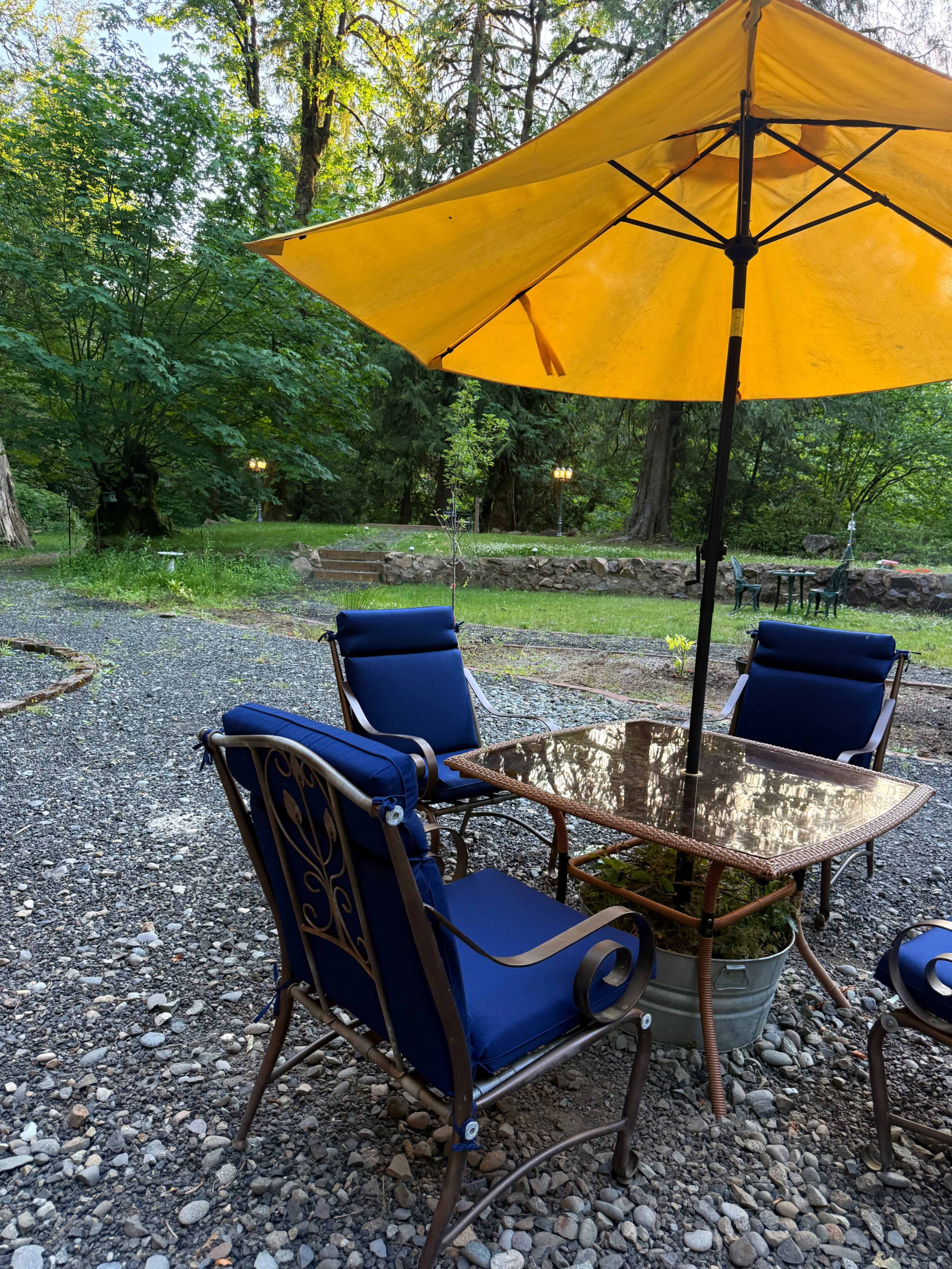 A patio set with blue chairs and a table is shaded by a yellow umbrella, surrounded by trees and gravel pathways.