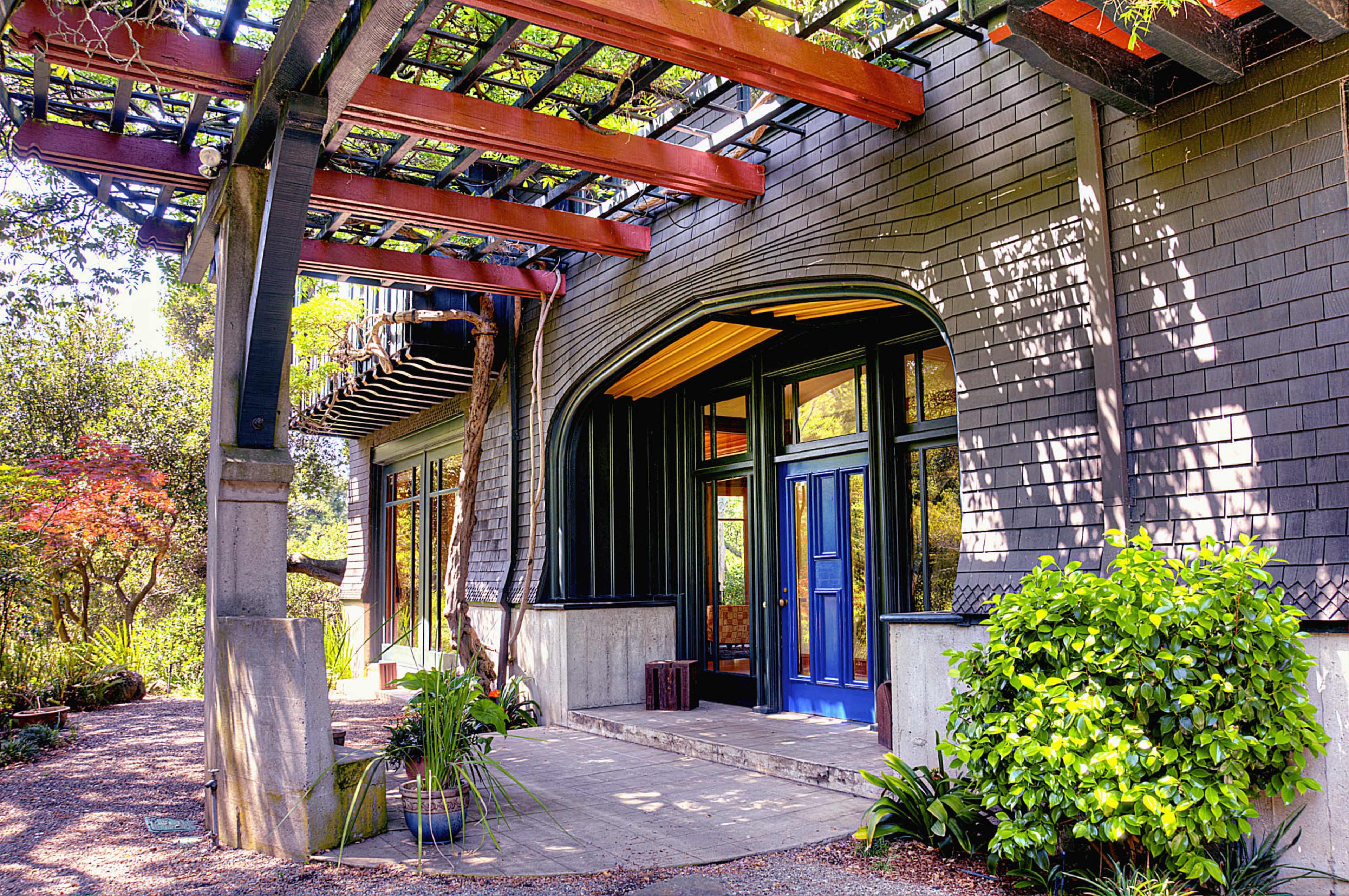 A modern house entrance with a pergola and a pathway leading to a double blue door, surrounded by greenery.