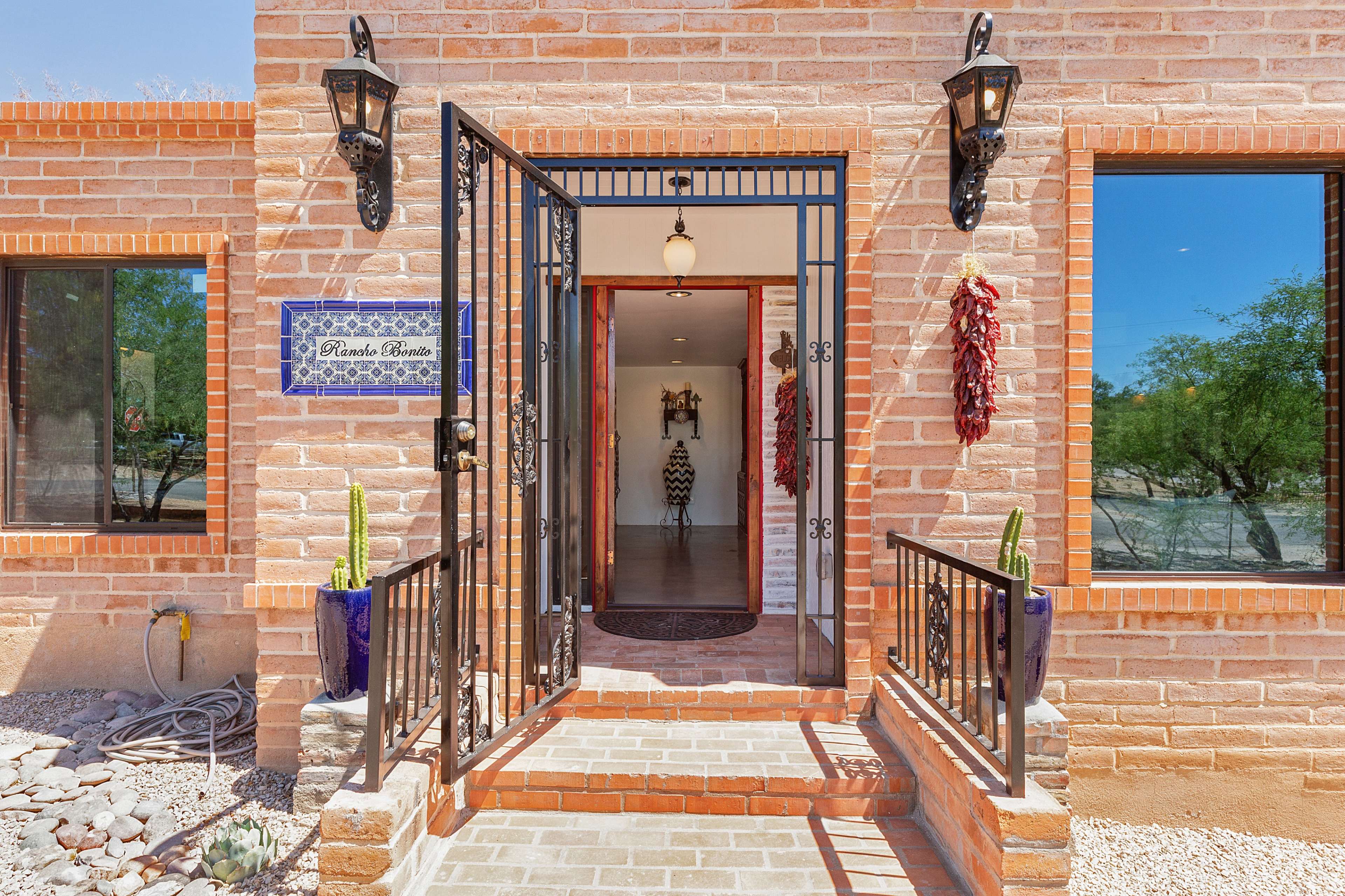 A brick building features an open wrought iron door with a welcome sign, leading to a well-lit interior flanked by large windows.