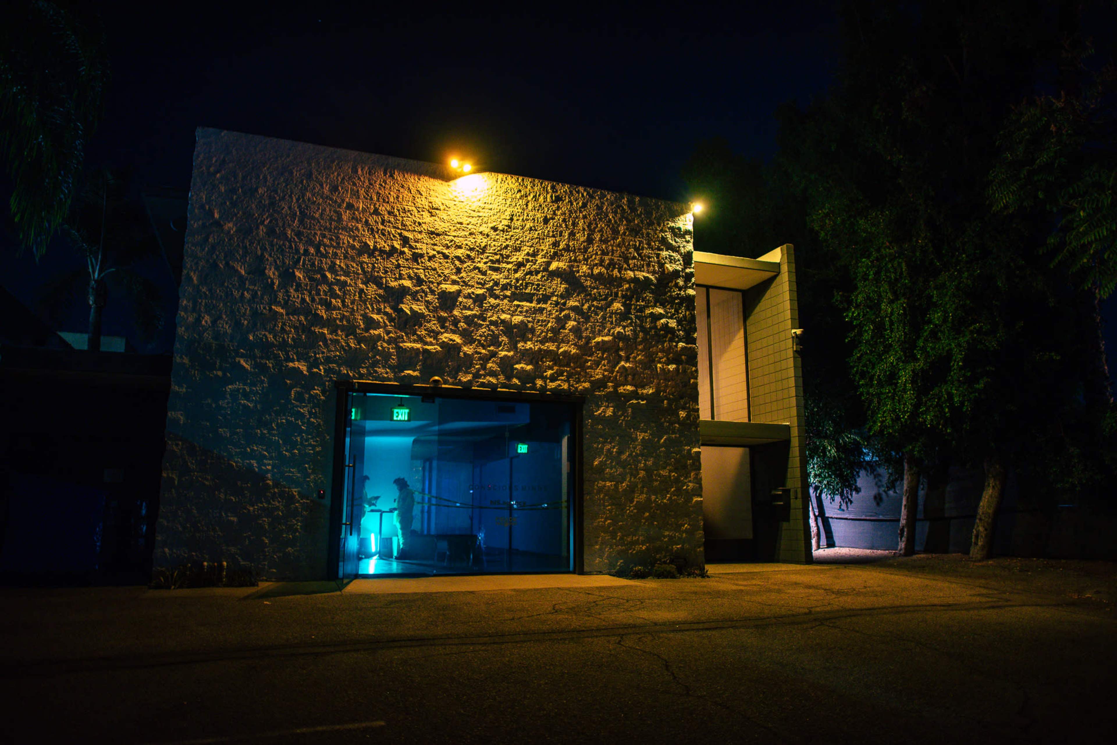 A modern building with a textured exterior is illuminated at night, featuring large glass doors that reflect blue light.