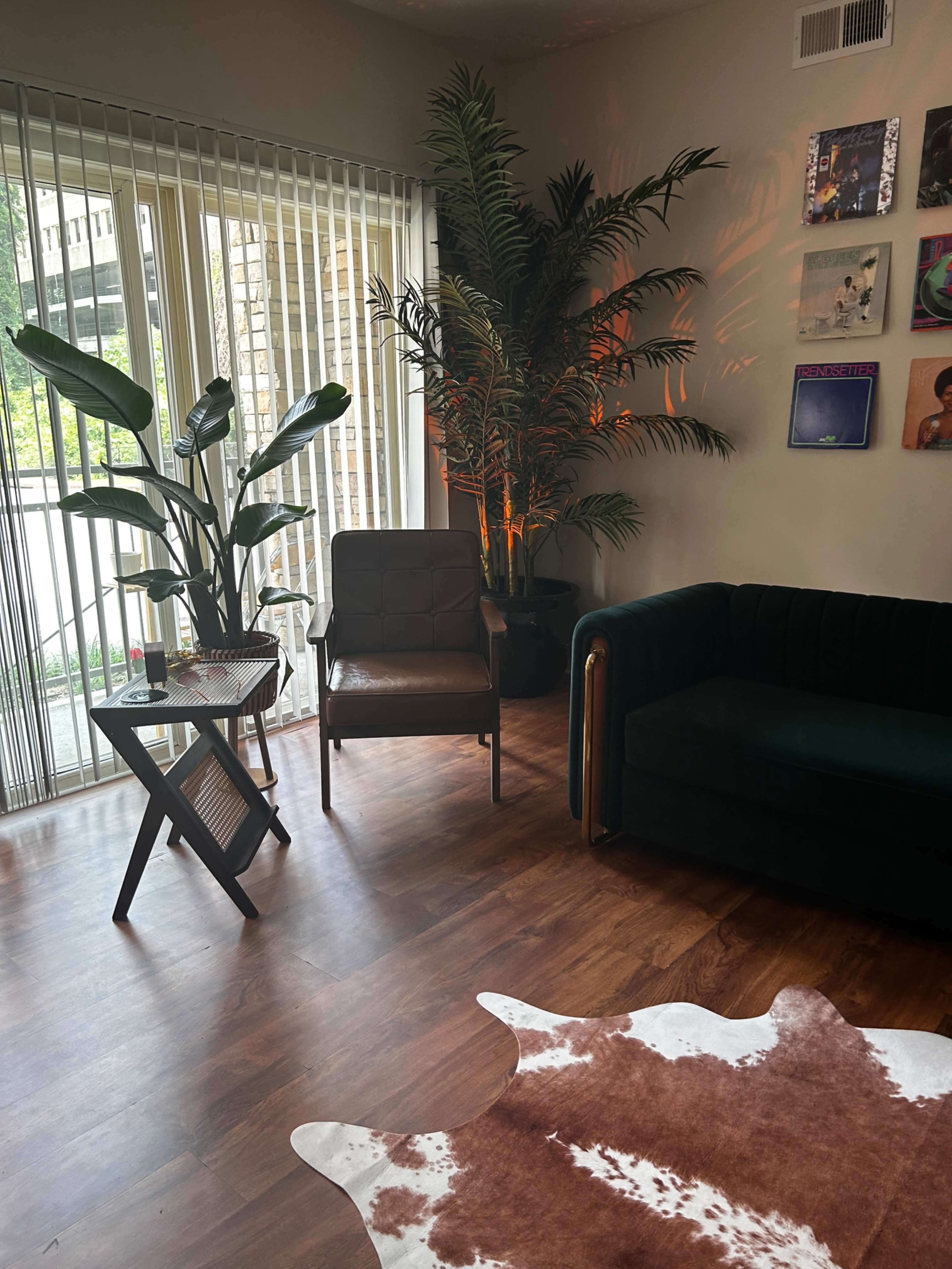 A living room featuring a green couch, a brown chair, a coffee table, and large indoor plants near a set of sliding glass doors.