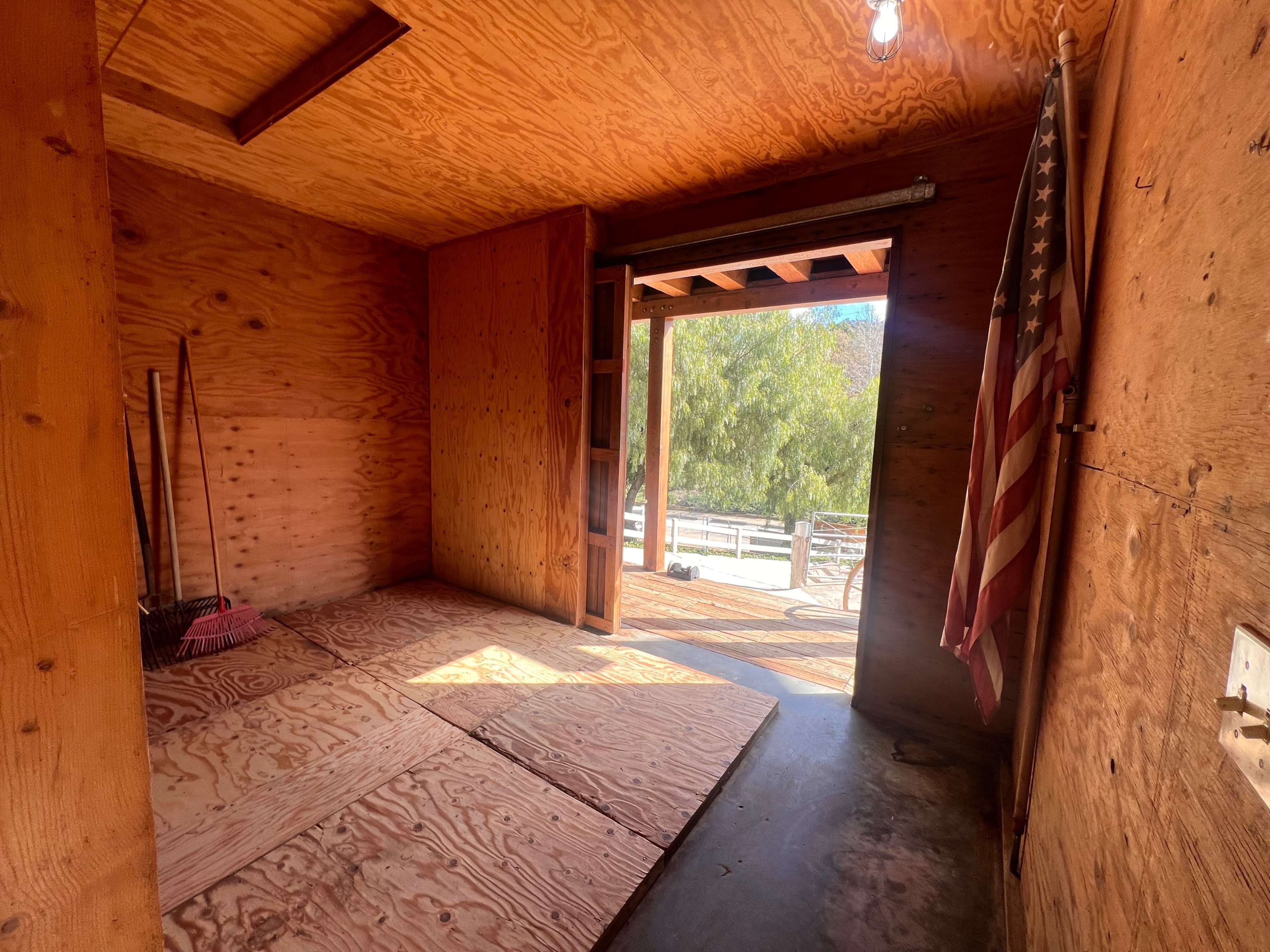 The interior of a wooden shed features plywood flooring, a broom leaning against the wall, and an open door leading to an outdoor area with an American flag visible.