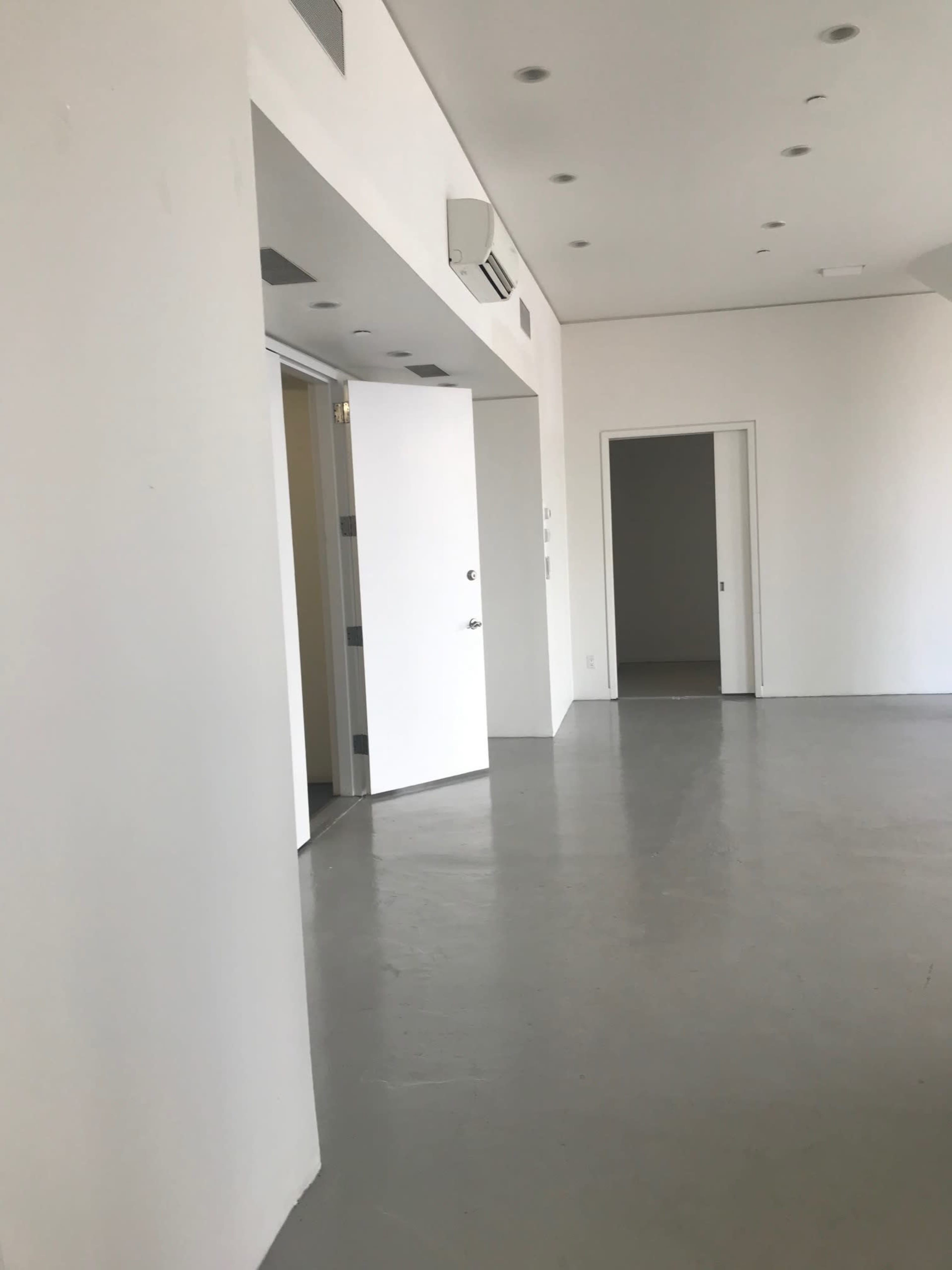 A spacious, empty interior with white walls and a gray floor, featuring several doors and ceiling lights.