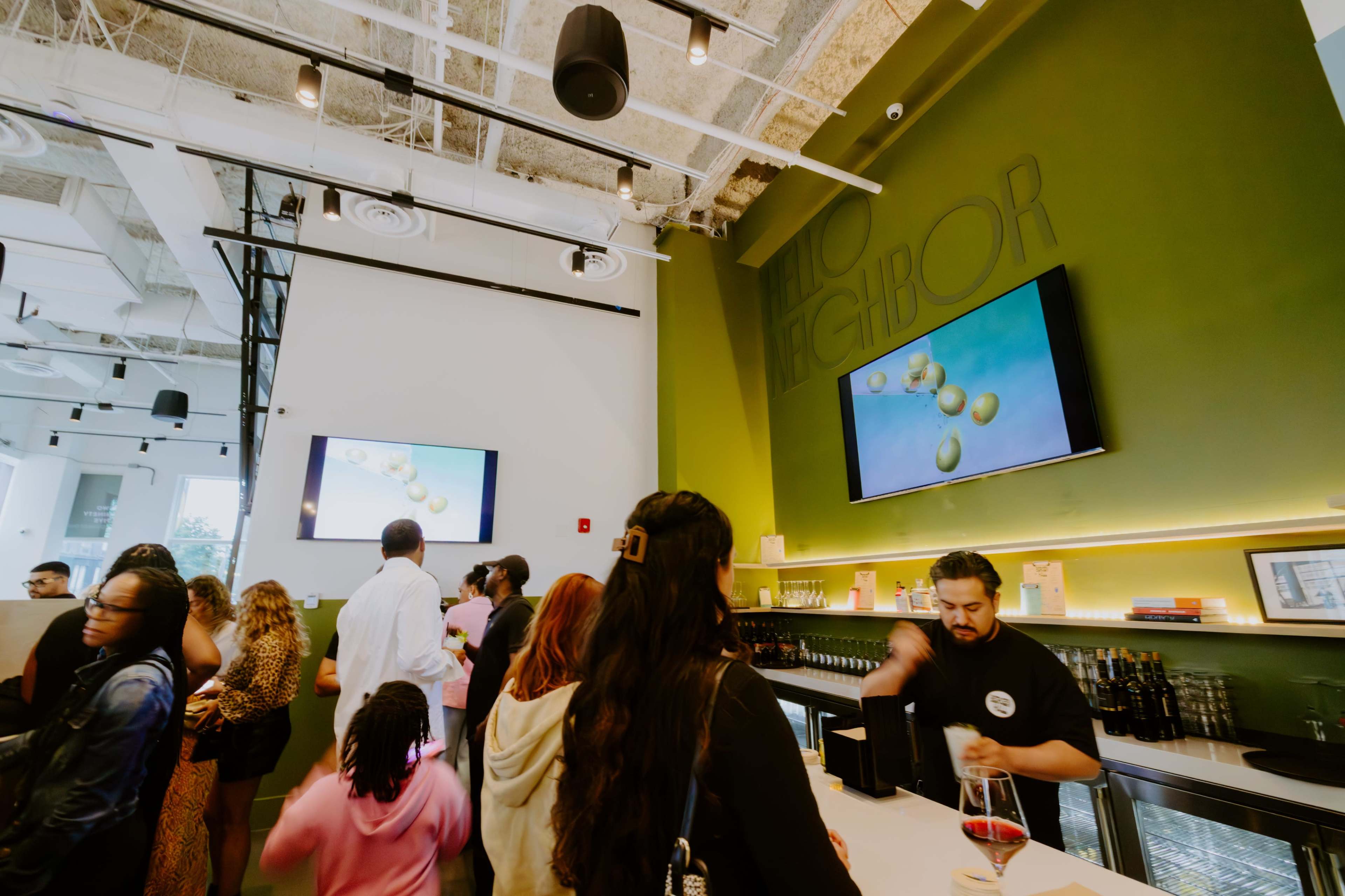A crowd of customers gathers in a modern café with green walls and two large screens displaying visual content.