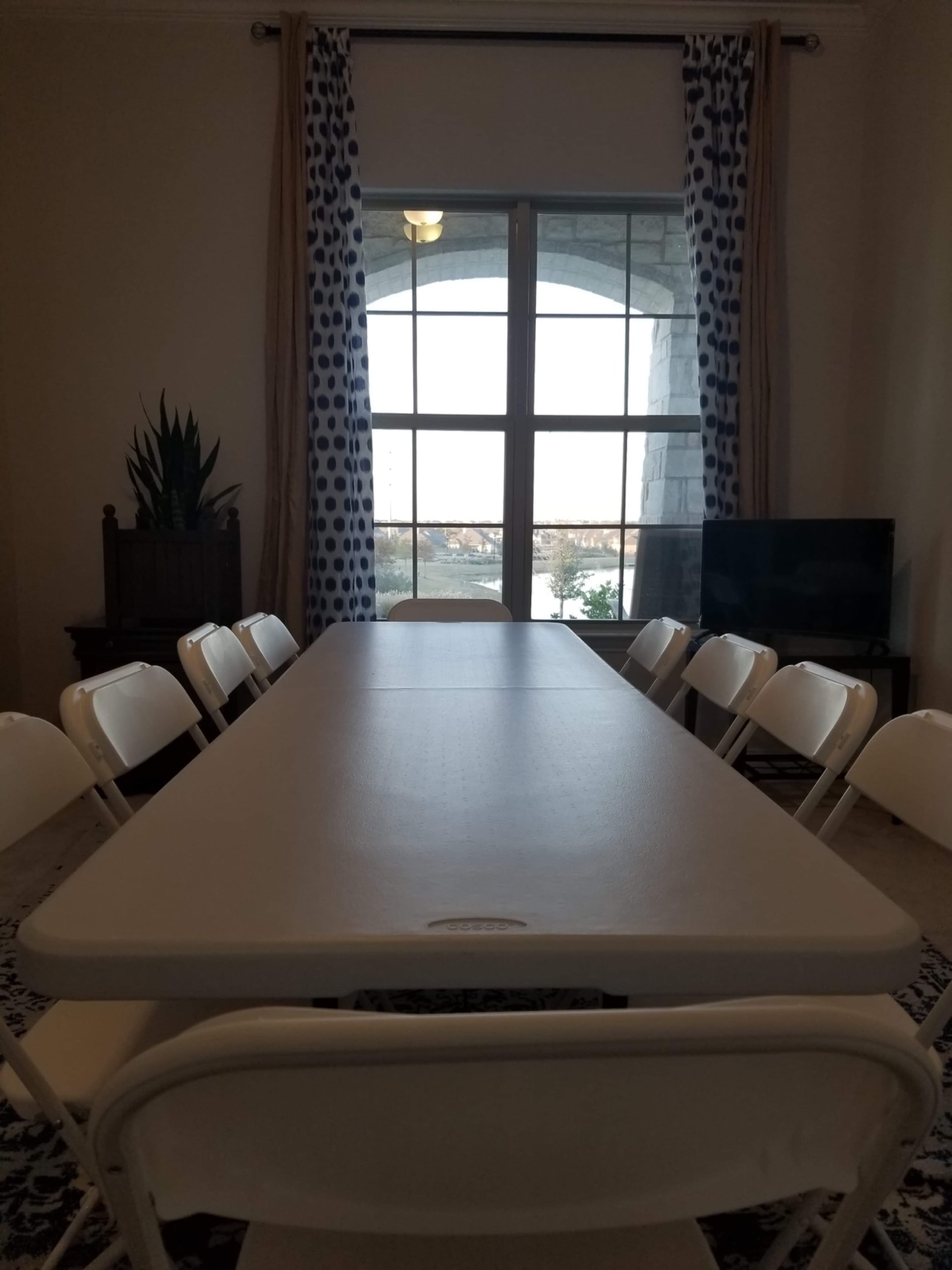 A long, rectangular table surrounded by folding chairs is set up in a room with large windows and a view of the outdoors.