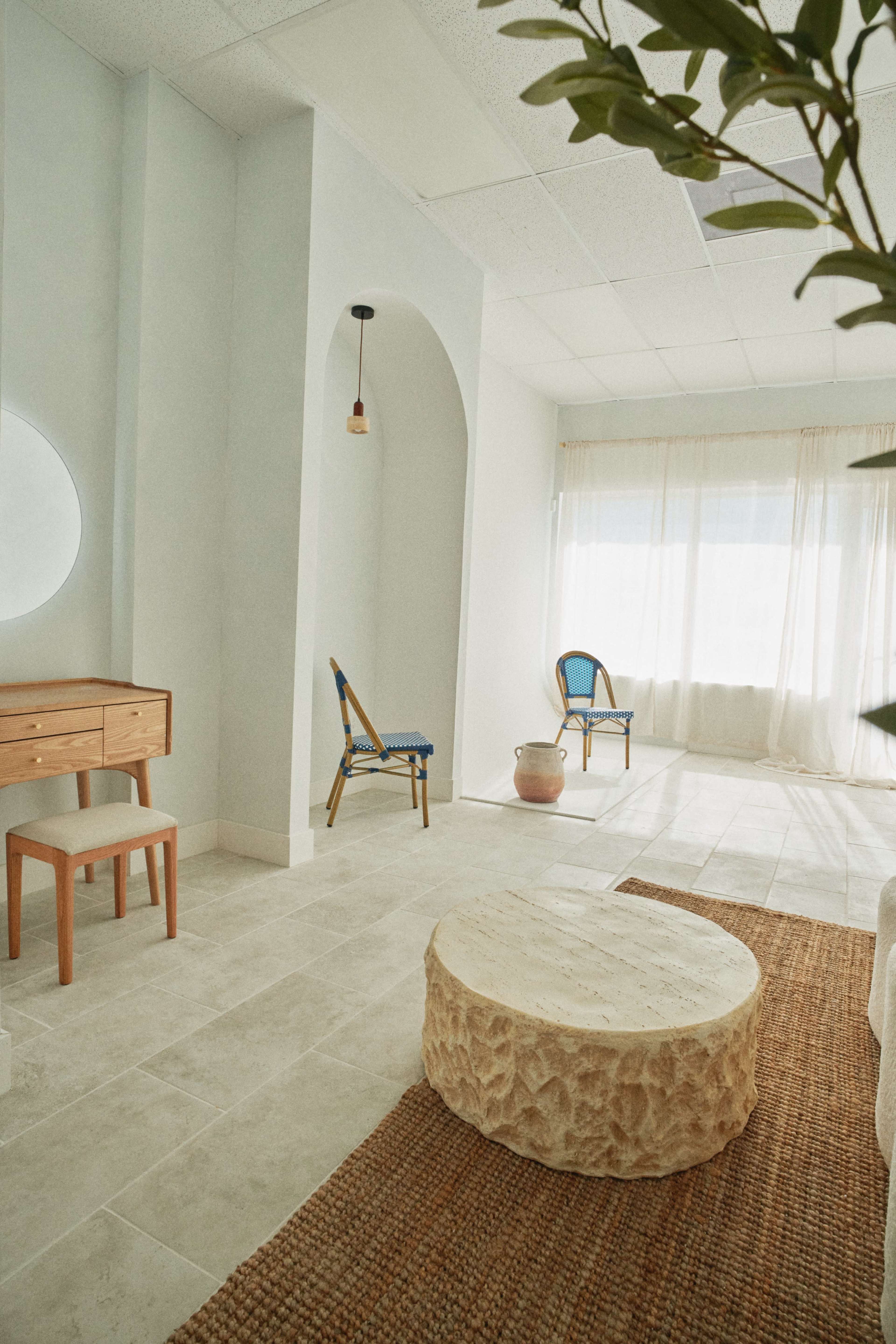 The image shows a light-filled room with a minimalist design, featuring a wooden vanity, a round stone coffee table, a chair, and large windows adorned with sheer curtains.