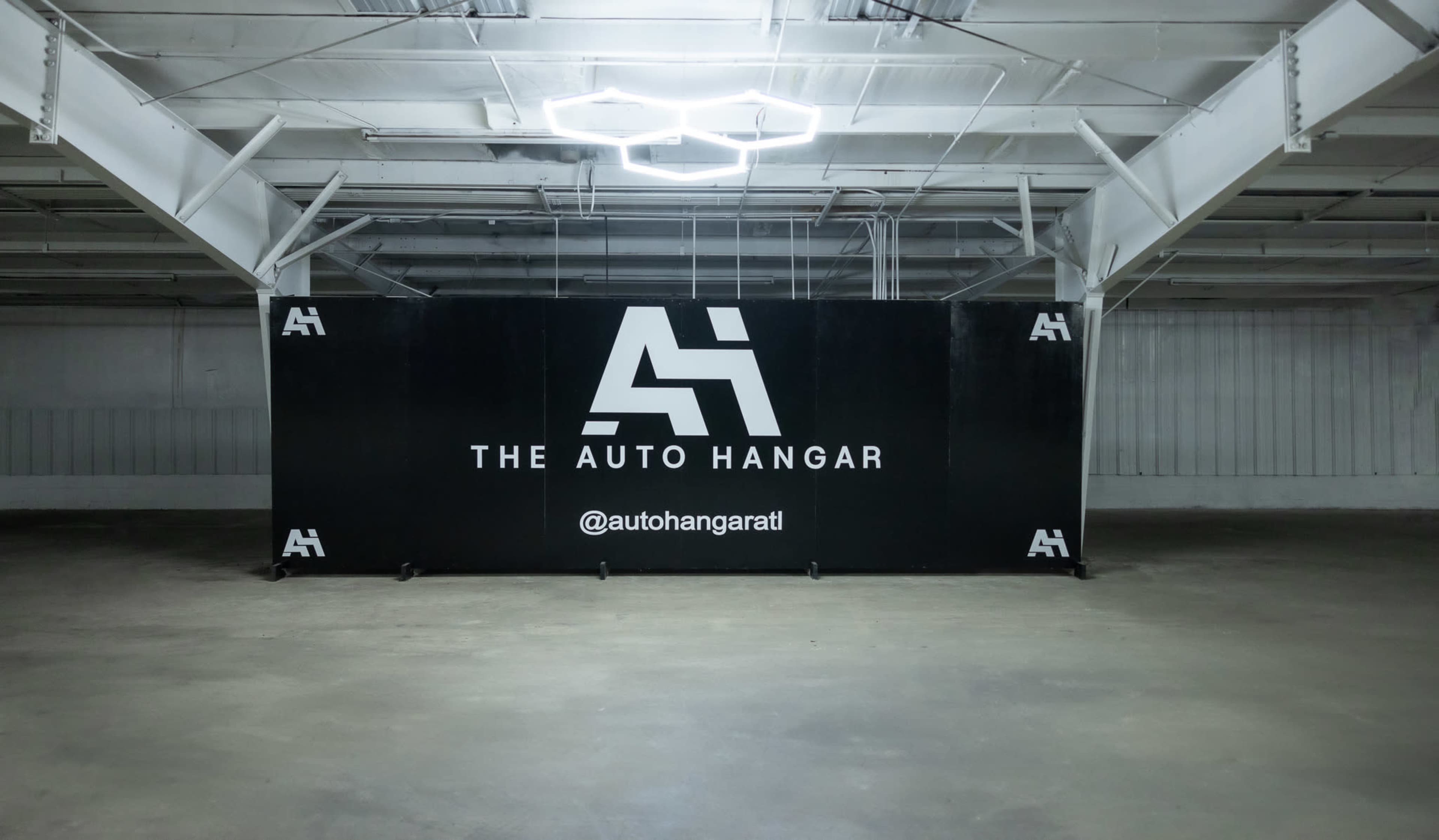 The image shows a black backdrop with the logo "THE AUTO HANGAR" and the Instagram handle "@autohangaratl" in an empty industrial space.