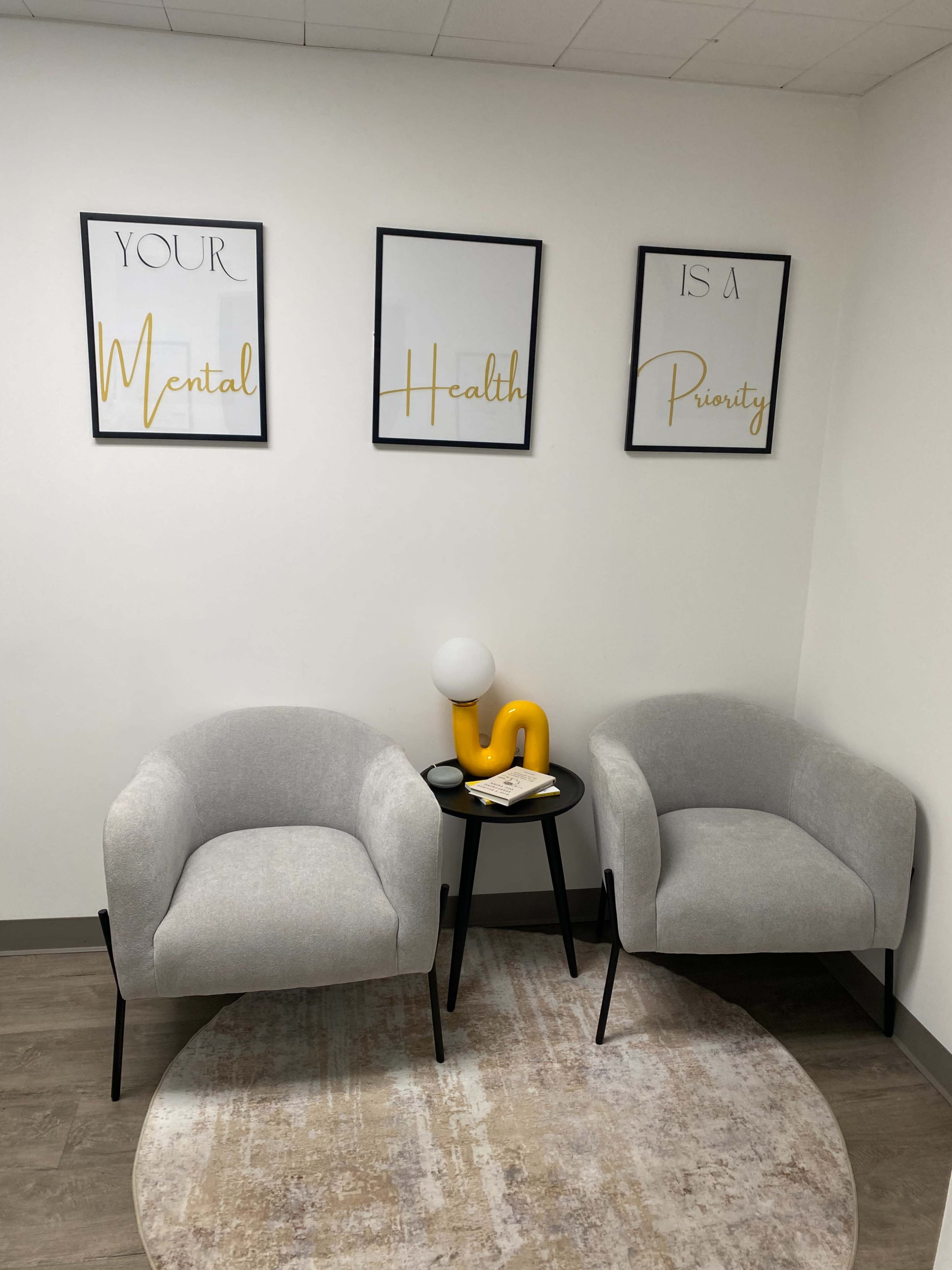 The image shows a small, inviting waiting area with two gray chairs, a round table, and framed motivational posters on the wall.