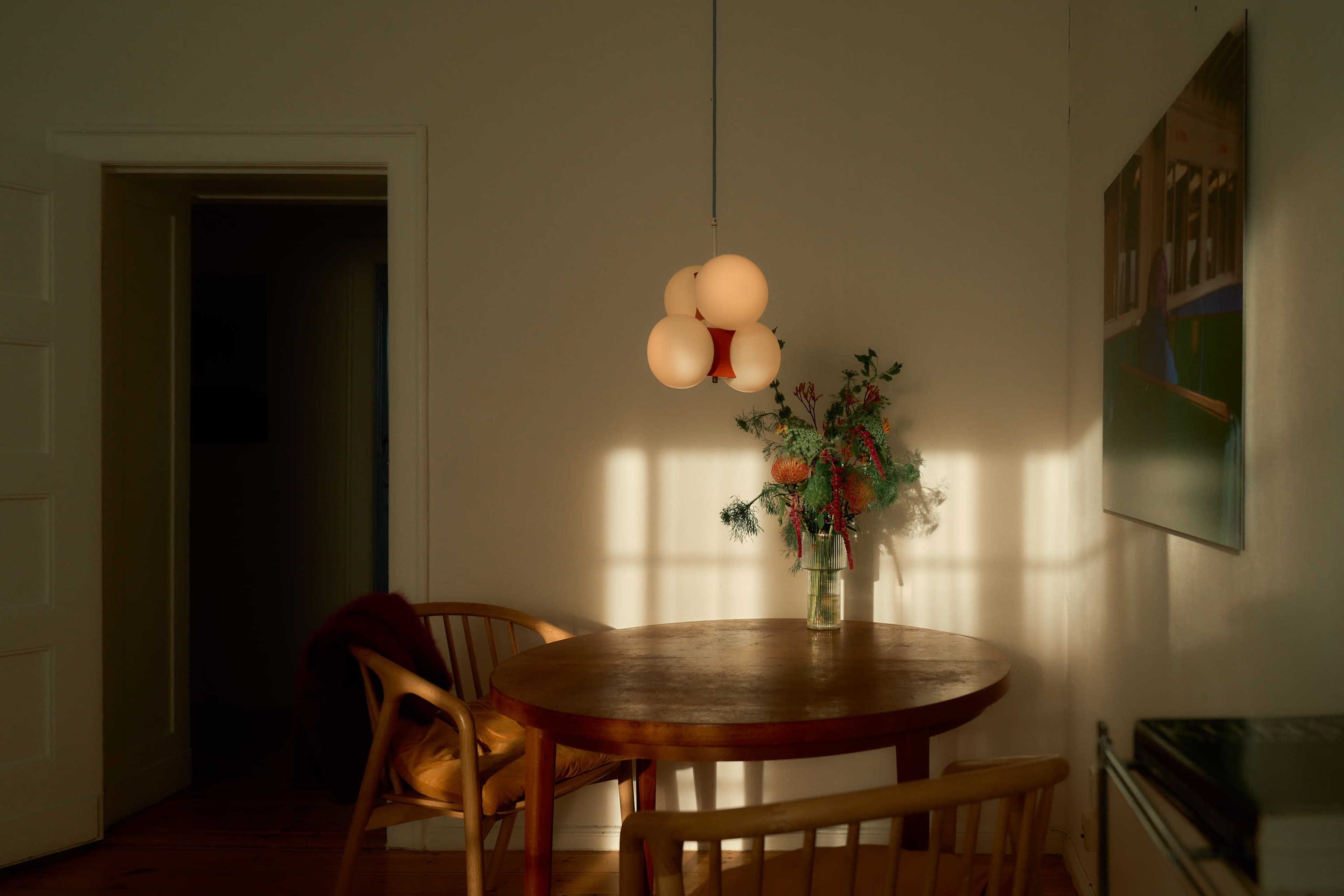 A round wooden table is set in a softly lit room, featuring a vase of flowers and a pendant light overhead.