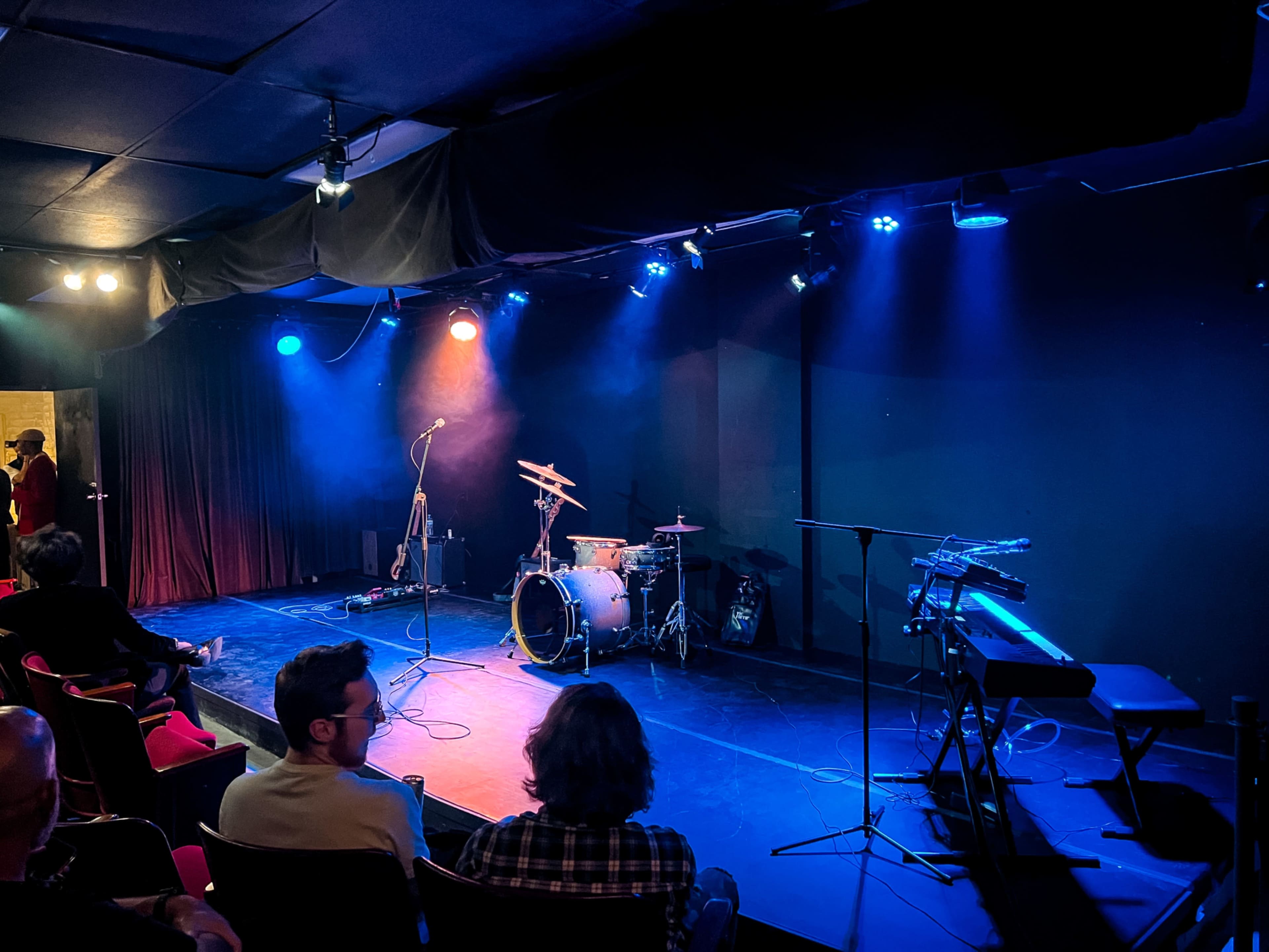 A stage is set up with musical instruments, including a drum set, keyboard, and microphone, under blue and orange stage lights in a small performance venue.
