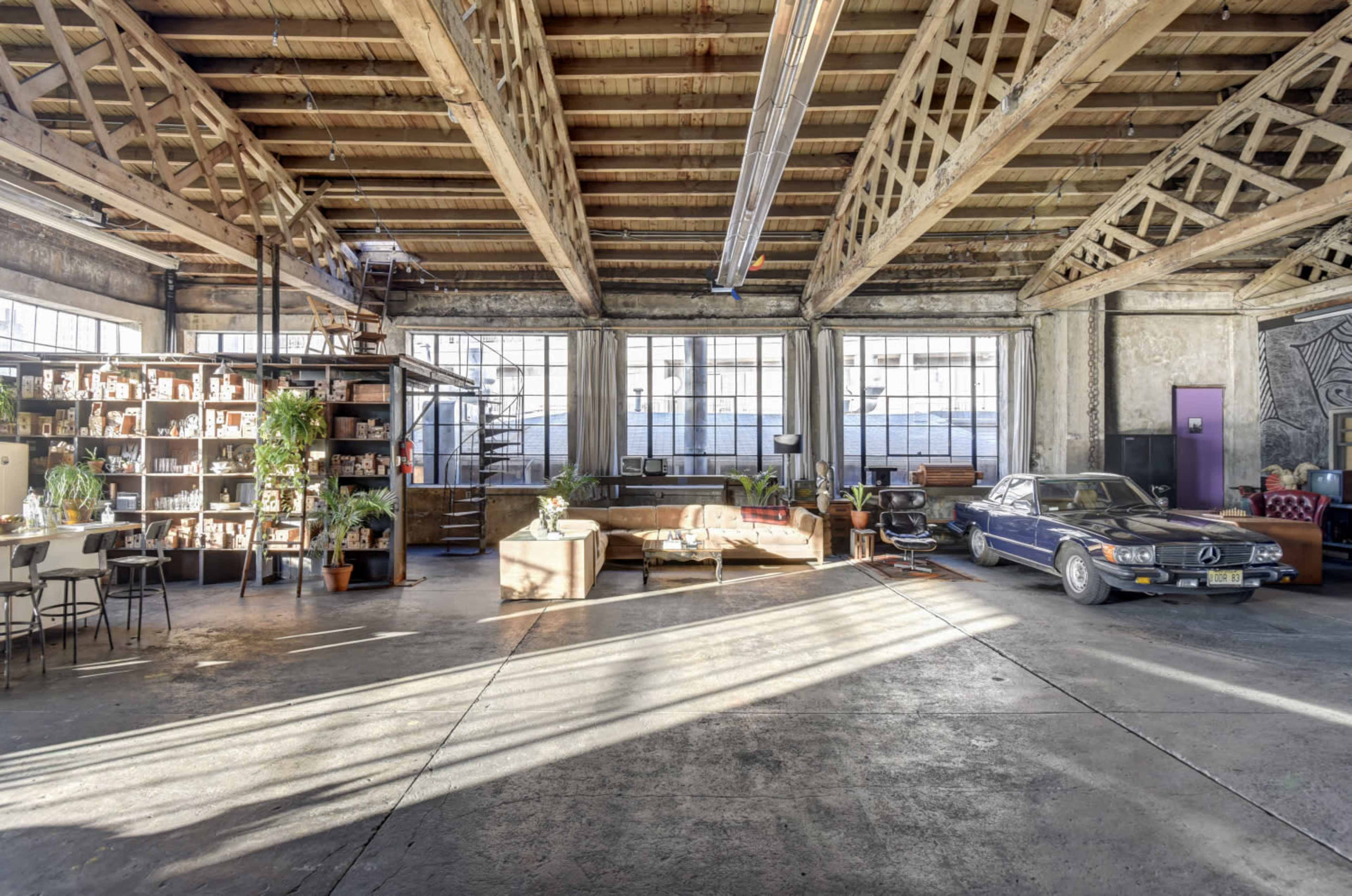 A spacious industrial loft with tall windows, wooden beams, and a mix of living and workspace areas, including a vintage car parked beside a seating area.