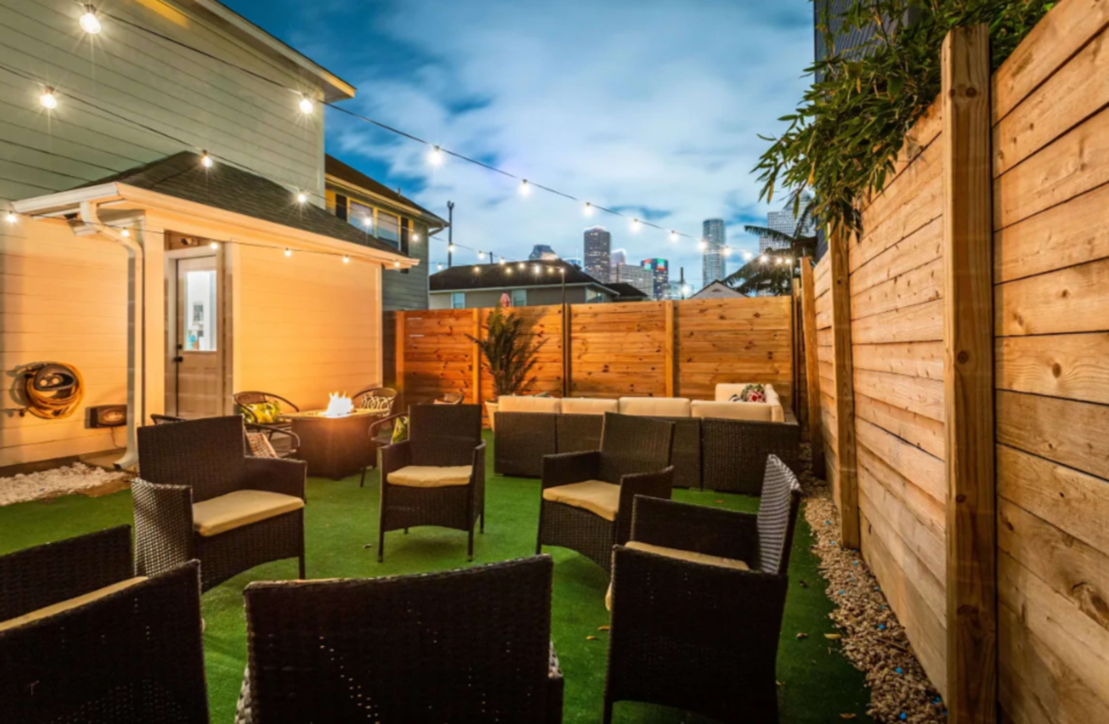 A cozy outdoor seating area with wicker chairs arranged around a central fire pit, set against a backdrop of a city skyline at dusk.