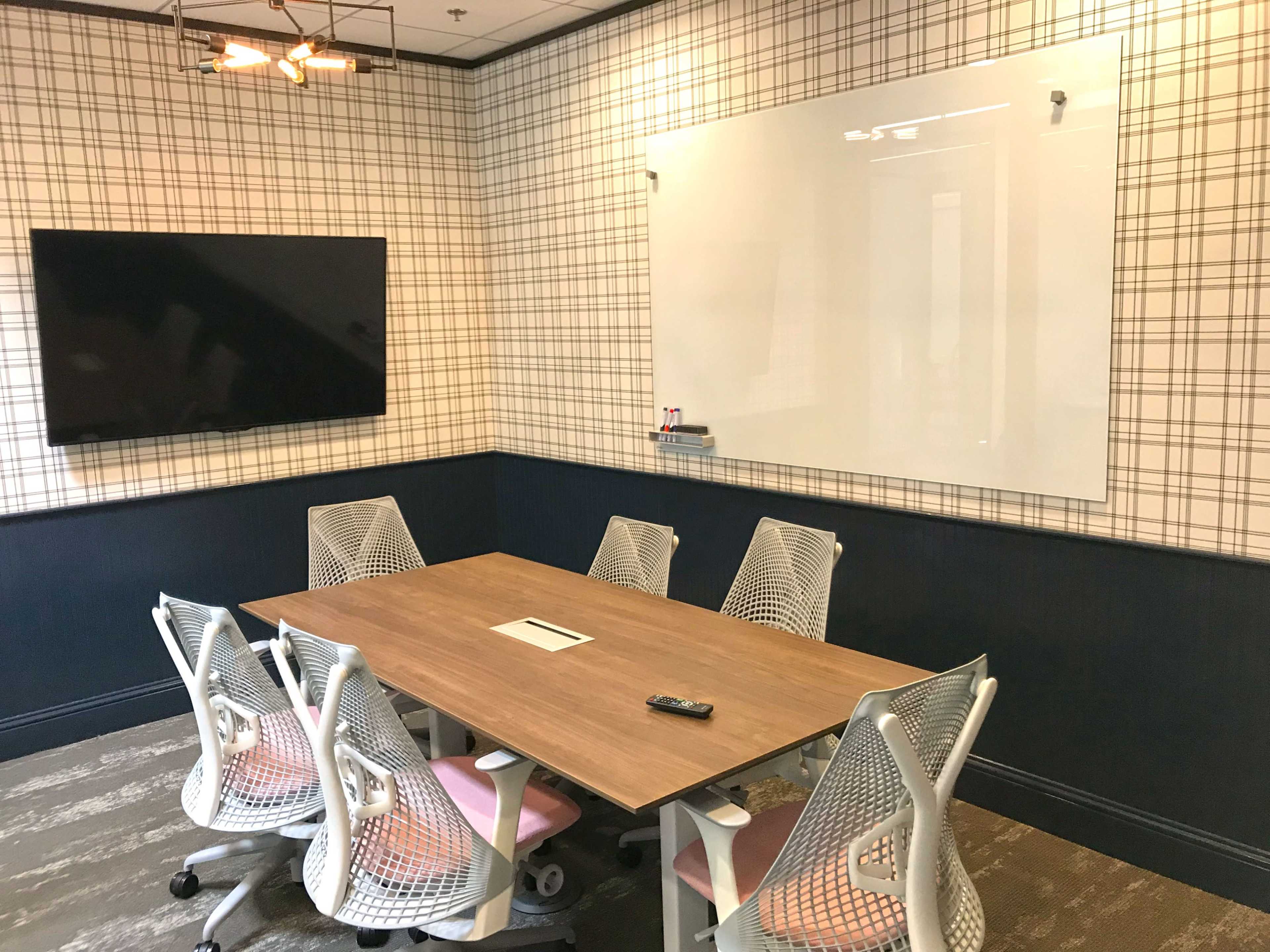 A meeting room with a large wooden table surrounded by six chairs, a wall-mounted TV, and a whiteboard on the patterned wallpaper.