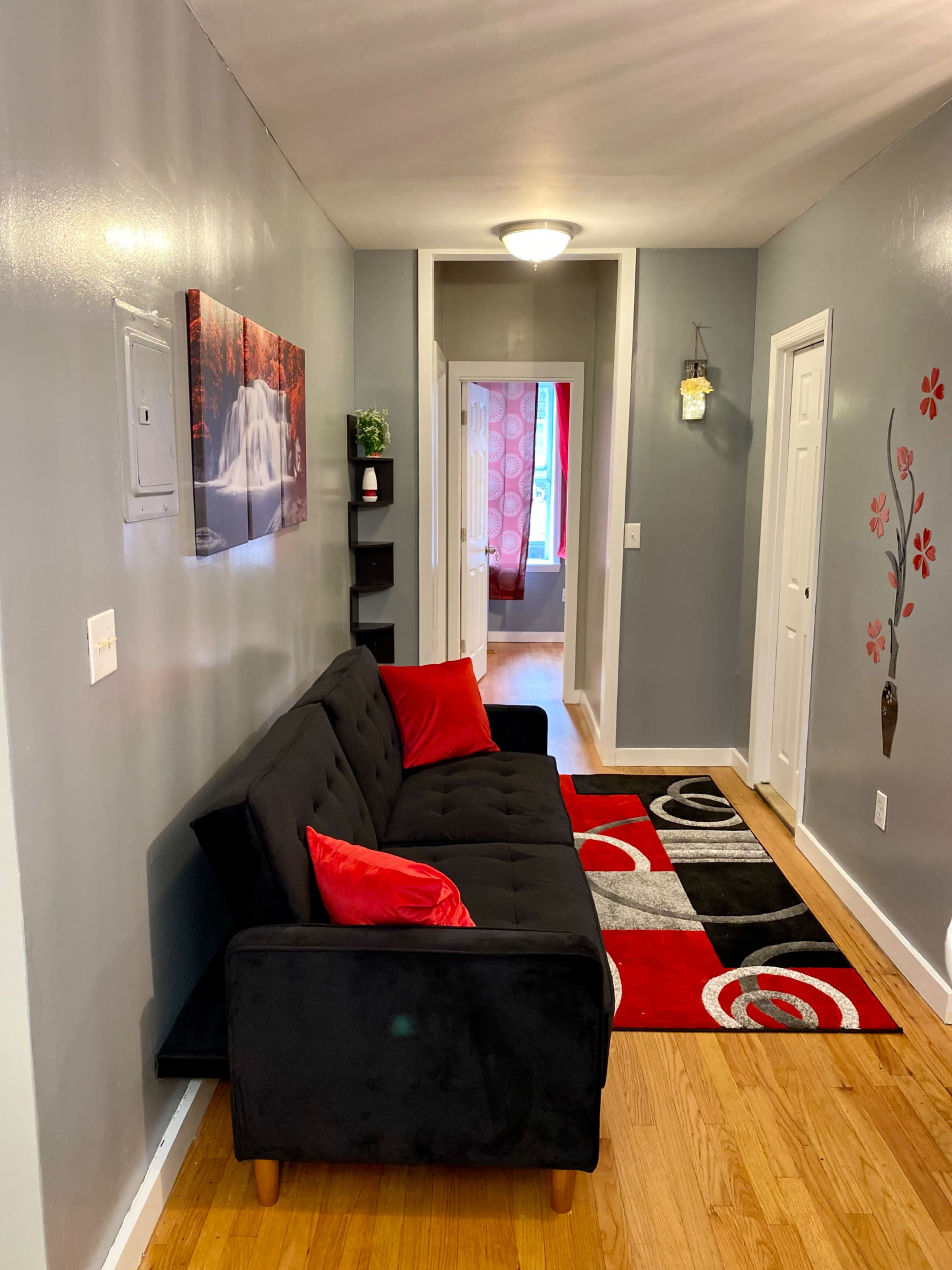 The image shows a narrow hallway with a black sofa, red accent pillows, a decorative rug, and a water-themed wall art, leading to a door at the end.